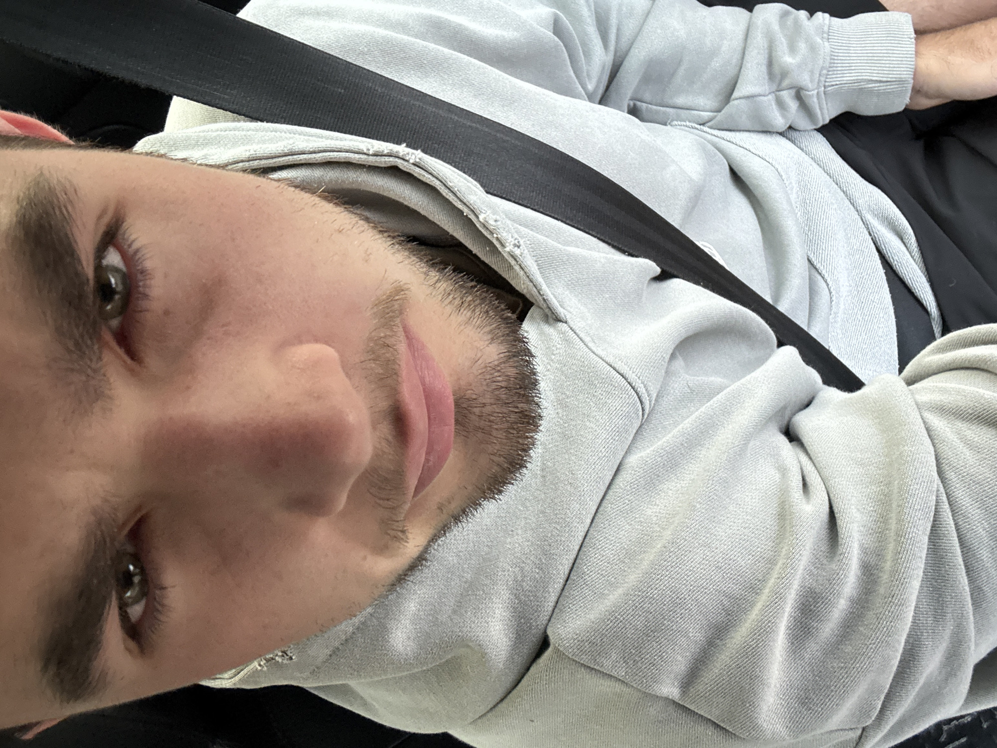 A close-up selfie of a young man with short, dark hair and a trimmed beard, wearing a light gray hoodie. He is wearing a black seatbelt. The location, San Bernardino Tlaxcalancingo, Mexico, is not directly discernible from the image.