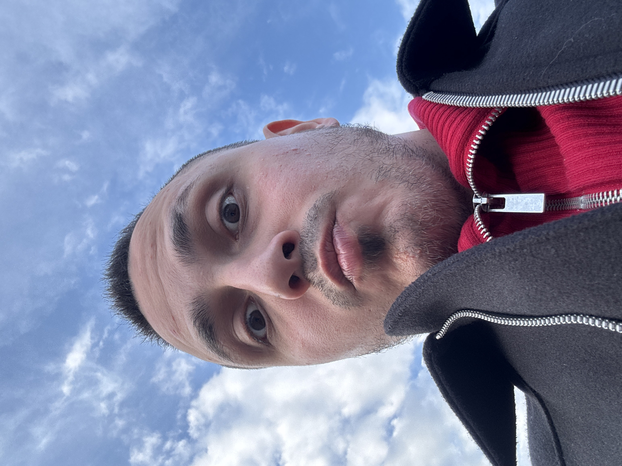 A man is photographed from a low angle, with his face close to the camera and the sky visible in the background. He has short, dark hair, light skin, and stubble. He is wearing a red zippered sweater underneath a black jacket. The zipper of the sweater is partially open. The background shows a cloudy sky, which suggests an outdoor location. There is no additional information suggesting the location of Saint Petersburg, Russia, therefore that cannot be confirmed.