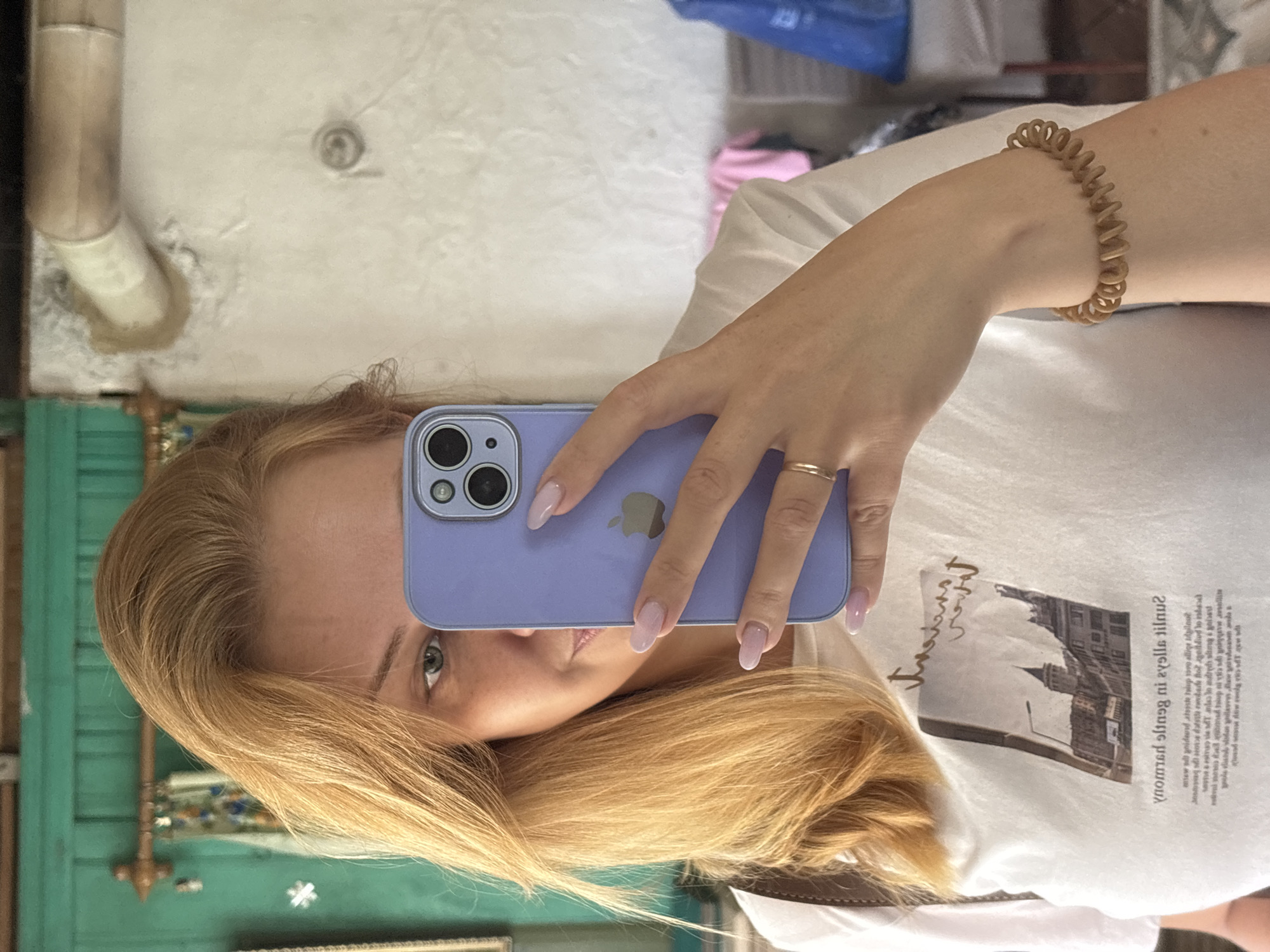 A Caucasian woman with blonde hair takes a selfie in a mirror. She is wearing a white t-shirt with a printed image and text. She holds a purple smartphone in her right hand, partially obscuring her face. Her left hand is visible, displaying light pink manicured nails and a gold ring. A brown coiled bracelet is worn on her left forearm. The background appears to be an indoor setting with a turquoise wooden structure, a white wall, and a hint of a window with curtains. The scene does not provide any specific information or context relating to the city of Cherkasovo, Russia.