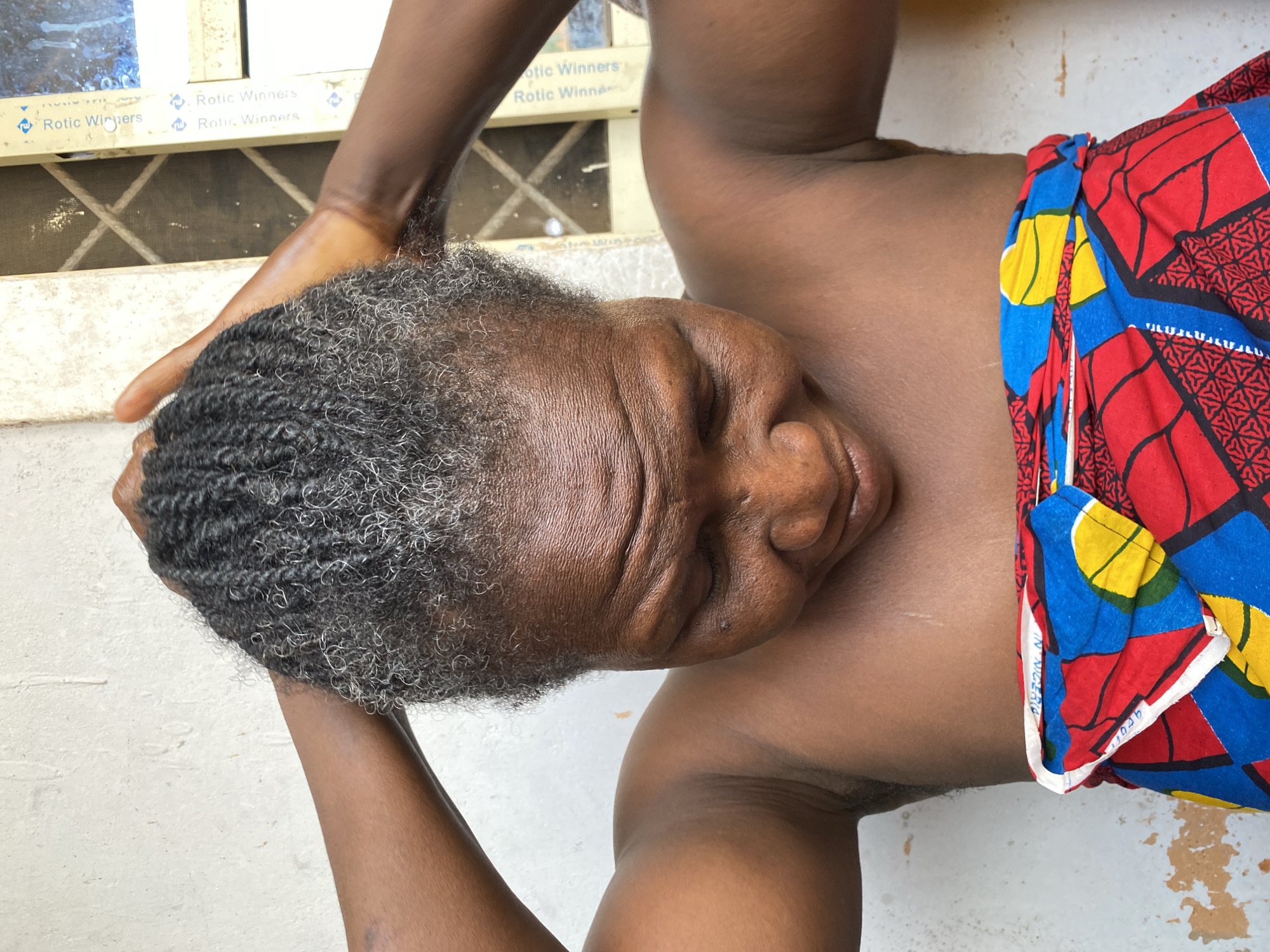 A close-up, tilted photograph depicts an older Black woman in Bukuru, Nigeria. Her head, featuring short, graying braided hair, is tilted towards the left of the frame. Her eyes are closed. Her face shows signs of aging. The woman's dark brown skin is visible on her neck, shoulders, and arms, and she is wearing a colorful garment with red, yellow, and blue patterns around her waist. Her left hand is positioned on the left side of her head. A window with an aluminum frame and a grid pattern is visible in the upper left corner of the shot. The background consists of a white wall.