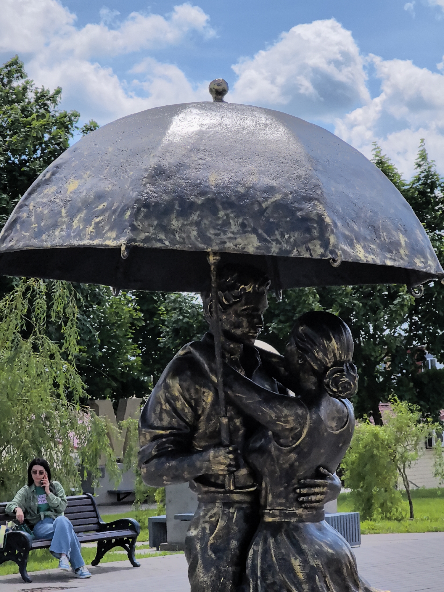 In the city of Unknown, Unknown, a large, weathered metal sculpture depicts a couple embracing under an umbrella. The umbrella, also made of metal, is open and positioned above the figures, casting them in shadow. The sculpture has a patinaed finish, with accents of gold paint visible on the edges and details of the figures and umbrella.
To the left of the sculpture, a young woman sits on a park bench. She is wearing a green collared shirt over a green t-shirt, blue jeans, and white sneakers. She has sunglasses on and is holding a mobile phone to her right ear, appearing to be in conversation. A green handbag rests on the bench beside her.
The environment is a park or outdoor public space. Lush green trees and foliage form a backdrop behind the sculpture and the seated woman. The sky above is blue with scattered white clouds. A large, stylized "M" is visible in the background, partially obscured by greenery, possibly a decorative element or part of a building. The ground is paved, with patches of grass.