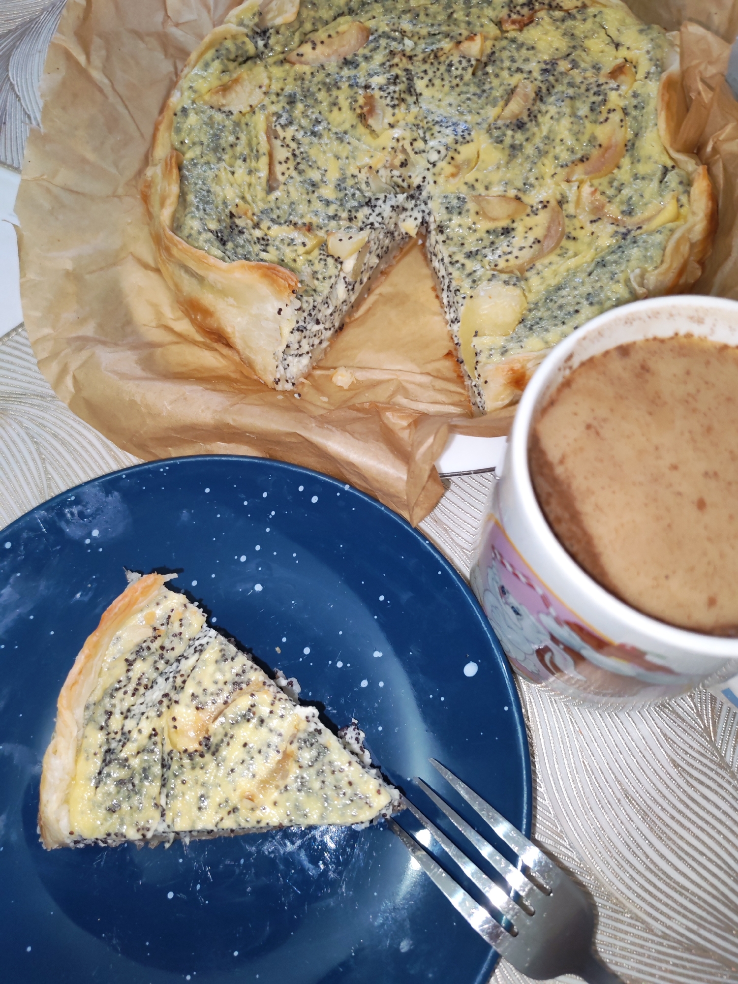A slice of savory pie with poppy seeds and what appear to be slivers of apple or pear sits on a dark blue plate with white speckles. A fork rests on the plate. In the background, a whole pie, partially sliced, is presented on brown parchment paper. To the right, a mug with a cartoon unicorn design holds a brown beverage, likely coffee or hot chocolate. The scene is set on a table with a textured, light-colored tablecloth. The overall lighting is dim. The image appears to be a still-life photograph of food and drink, taken indoors.
