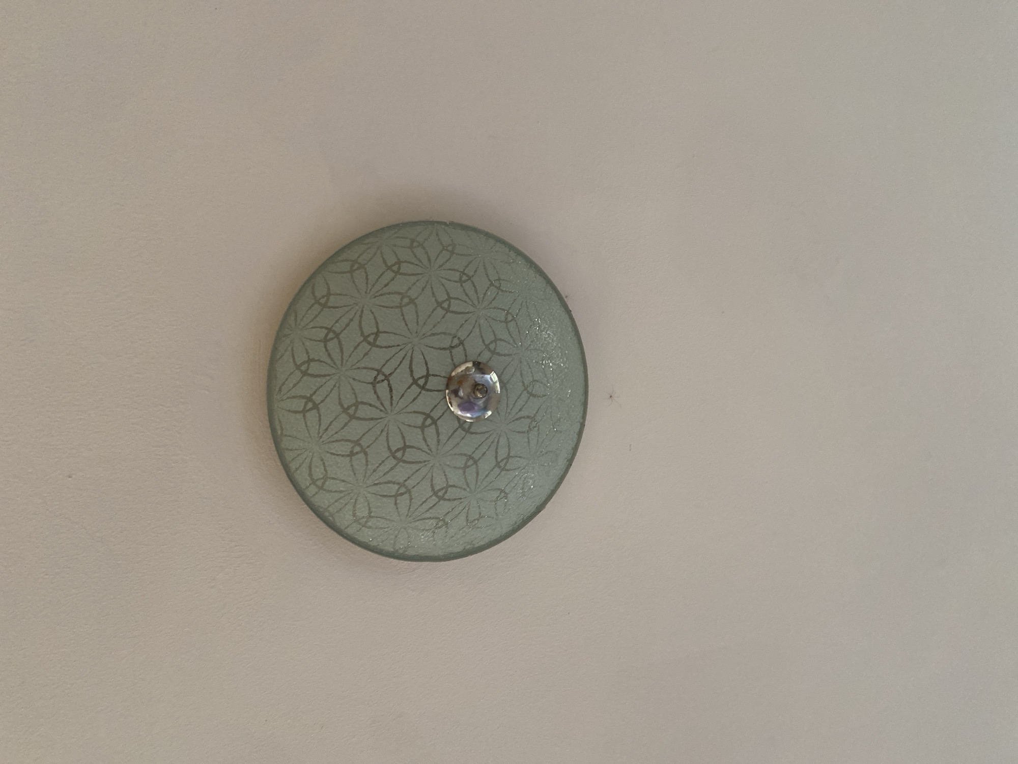 The image shows a circular ceiling light fixture affixed to a light beige ceiling. The fixture has a light green, textured glass cover with a floral pattern. A chrome-colored knob or screw is located in the center of the glass cover. The background is a plain, light beige ceiling. The location in the city of Ado, Nigeria is not identifiable from the image.