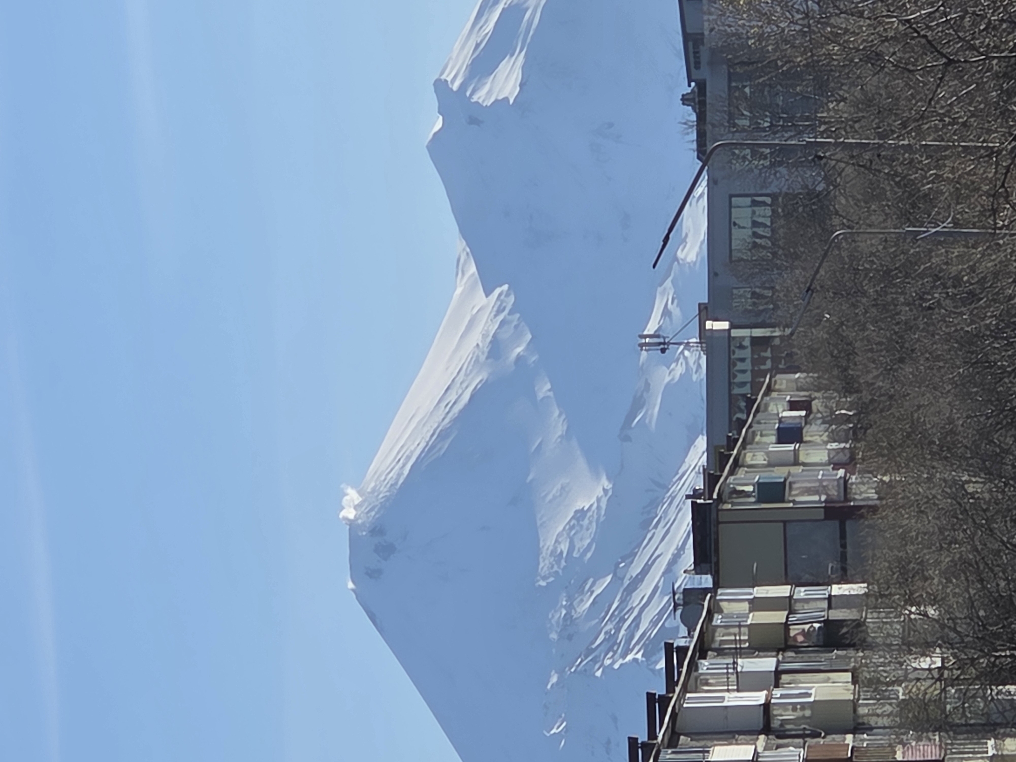The image shows a snow-covered mountain rising behind a building in the city of Unknown, Unknown. The building is tall and narrow, with many small windows and balconies. There are trees to the right of the building, which are leafless. The sky is clear and blue.