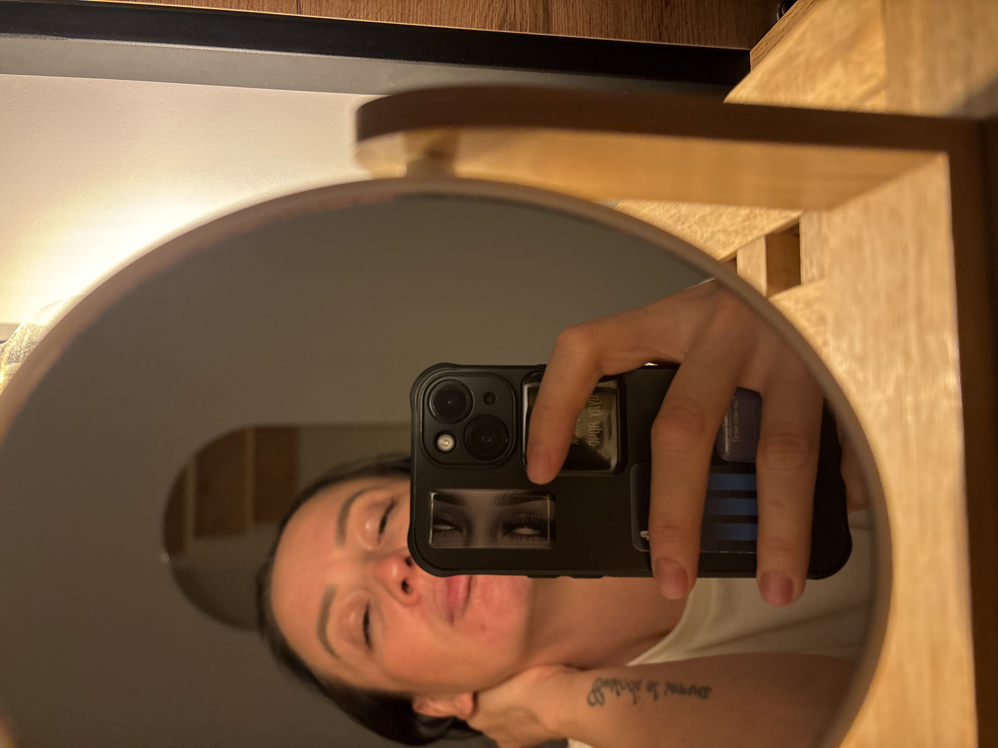 A selfie taken in a mirror. The mirror is round with a wooden frame. The person holding the phone has dark hair and a tattoo on their forearm. The phone is black and has multiple cameras. It also has a sticker of eyes on the back. The background appears to be a room with light-colored walls. There is no evidence indicating this photo was taken in Chaikovsky, Russia.
