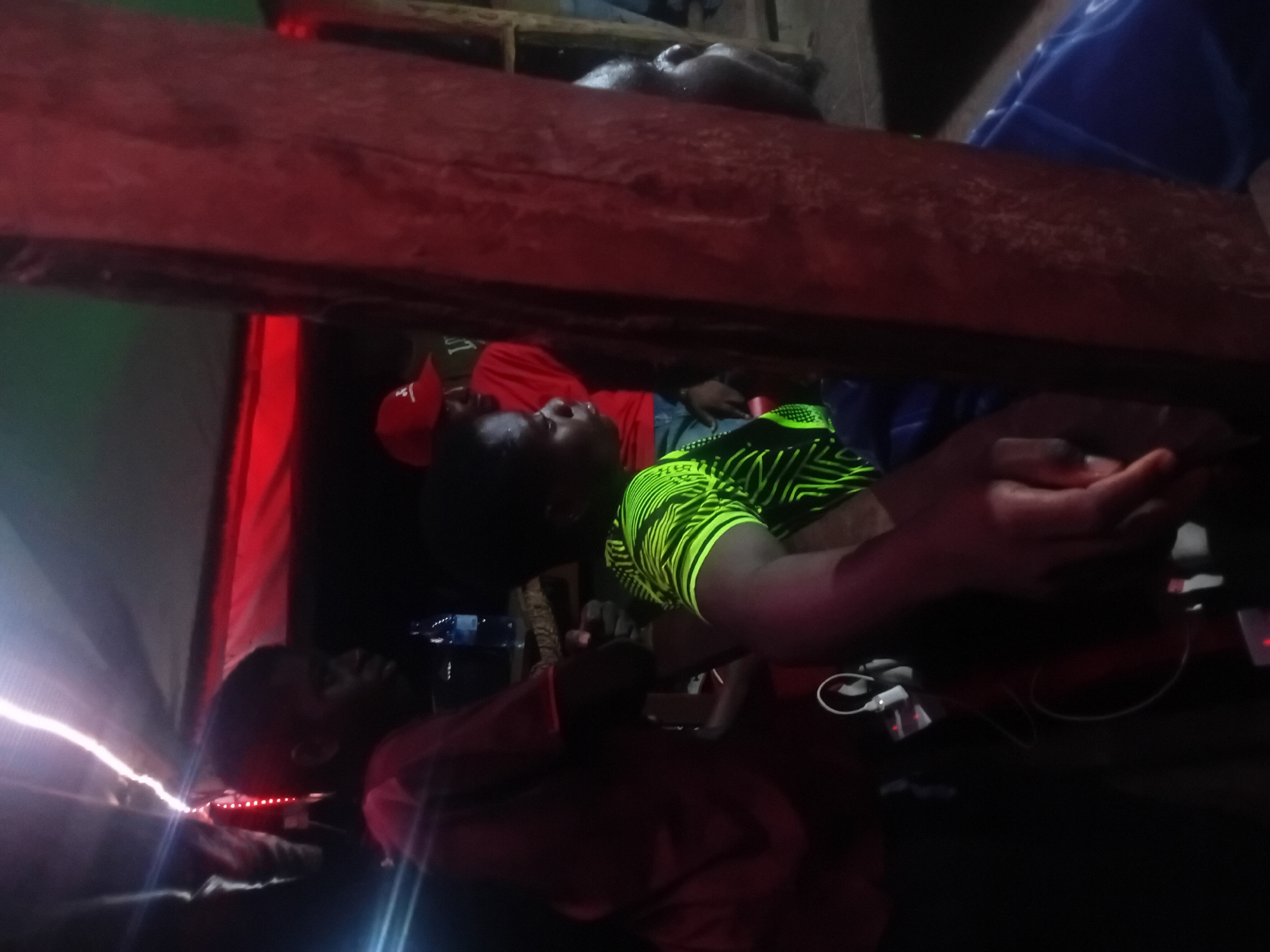 The image captures a dimly lit indoor scene, likely at night or in a poorly lit shelter, in Lokoja, Nigeria. The composition is vertical, showing several individuals resting within what appears to be a multi-level sleeping arrangement, possibly bunk beds or cots.
In the upper section, a wooden beam separates the levels. On the top level, part of a person's head and shoulder are visible, along with blue fabric, possibly a bag or blanket. Below the wooden beam, three individuals are on a lower level.
To the left, a person is wearing a red shirt and a red cap with faint white text, possibly "LI," visible on its front. This individual is lying down and looking upwards. Next to them, in the center, another person is seen in a bright green and black patterned short-sleeved shirt, also appearing to be lying down or reclining, looking towards the left. Their hands are near several electronic devices, including a white adapter/charger and cables. A faint red light emanates from a device nearby.
Further to the left, a fourth person in a darker red or brown shirt is partially visible, facing towards the left, with their face illuminated by a bright white light source just outside the frame on the far left. This light creates a strong glare. Below them, a small black device with red indicator lights is visible. The overall lighting includes strong reds, suggesting either decorative lighting or reflections from a red light source.
The scene conveys a sense of communal resting or winding down, possibly in a camp, dorm, or temporary lodging, with individuals using and charging personal electronic devices.