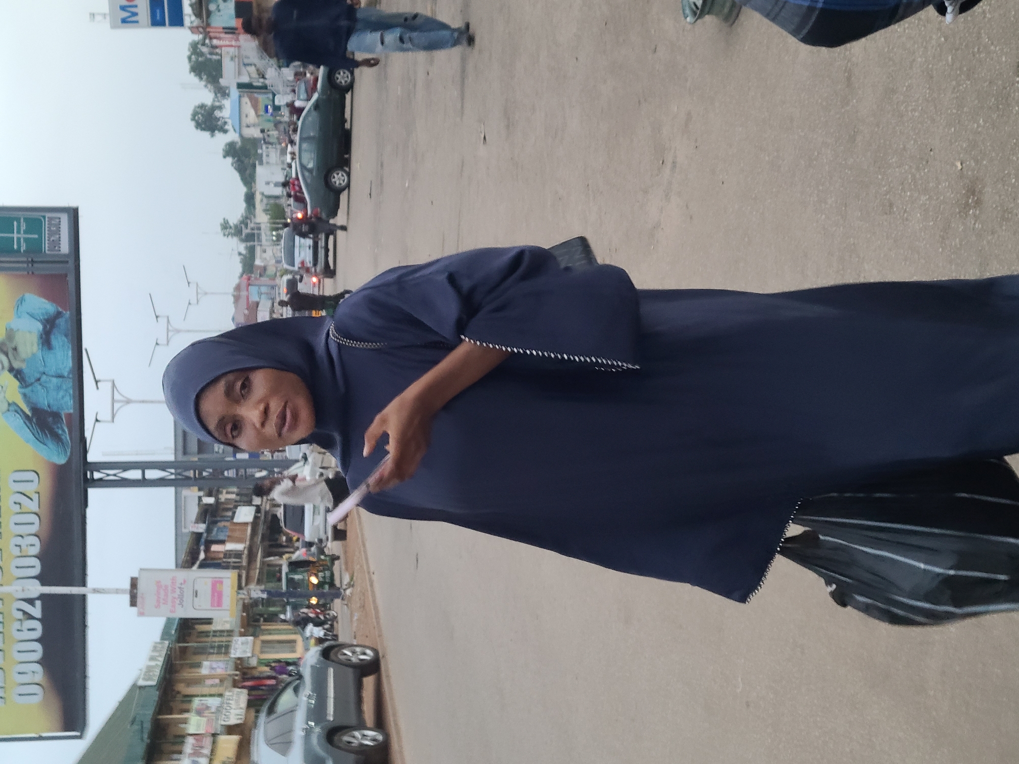 The image depicts a street scene in Jos, Nigeria. In the foreground, a woman wearing a dark blue hijab and dress is holding a smartphone in her right hand and a striped black plastic bag in her left. Her dress has decorative stitching along the cuffs. The background reveals a busy street with various vehicles, including a grey SUV on the left, a green sedan, and what appears to be a tuk-tuk. Buildings line the street, some with signage, suggesting a commercial area. Billboards are visible overhead. Other pedestrians are also present, including a person in ripped blue jeans walking away from the camera on the right. The overall atmosphere is that of a moderately trafficked urban environment during daylight hours, possibly overcast given the lighting.