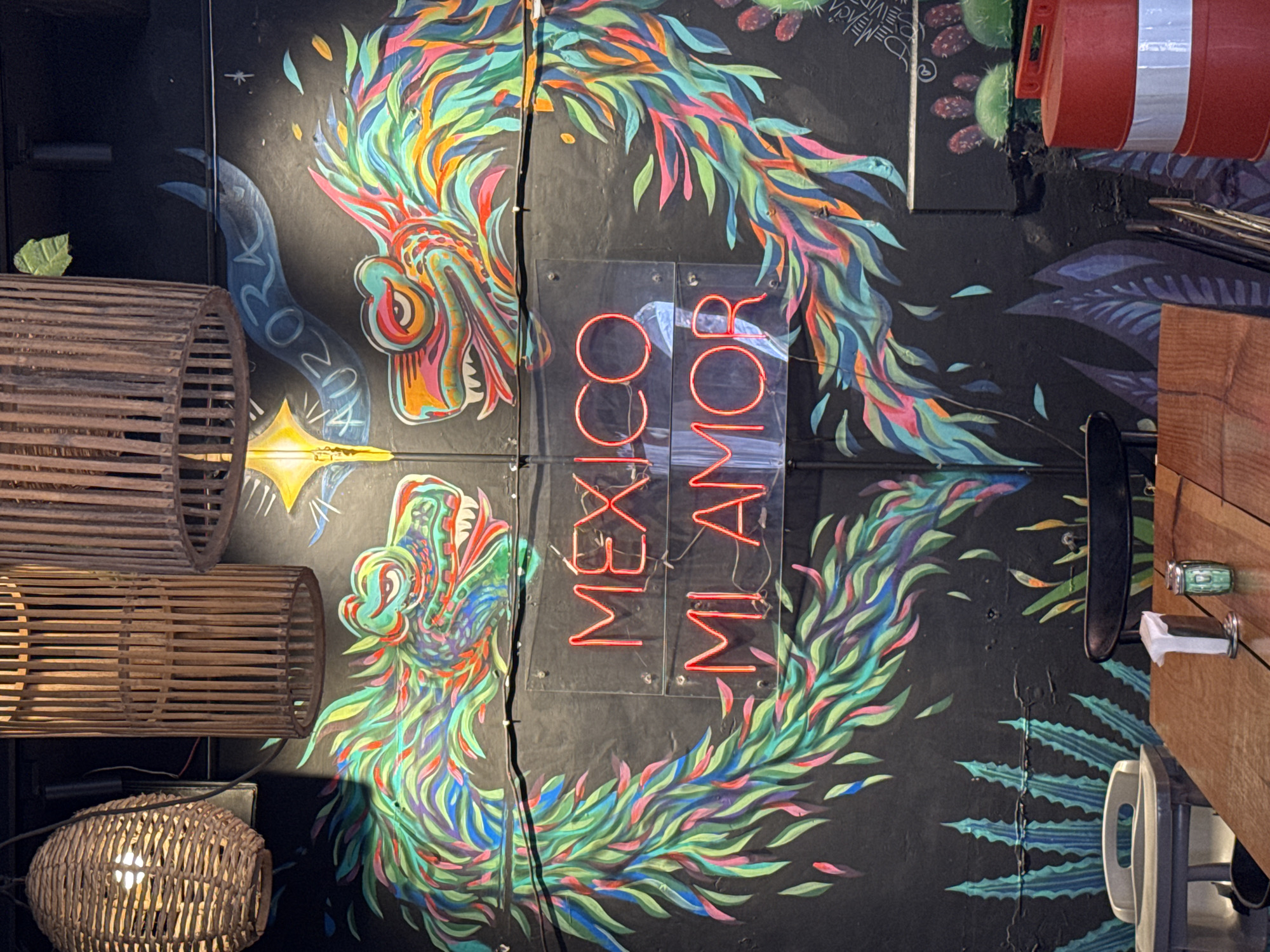 The photograph shows a wall in what is possibly a restaurant in Querétaro, Mexico. The wall is decorated with a colorful mural of two intertwined serpent-like creatures with vibrant, feather-like scales and ornate heads. Between the two creatures is a neon sign that reads "Mexico Mi Amor" vertically. To the left of the mural are three woven, cylindrical light fixtures, two long and one round. The environment is dimly lit, casting shadows that add depth to the scene. To the right, there is a partial view of a wooden countertop with a metal fixture. The overall impression is a blend of cultural art and contemporary design elements.