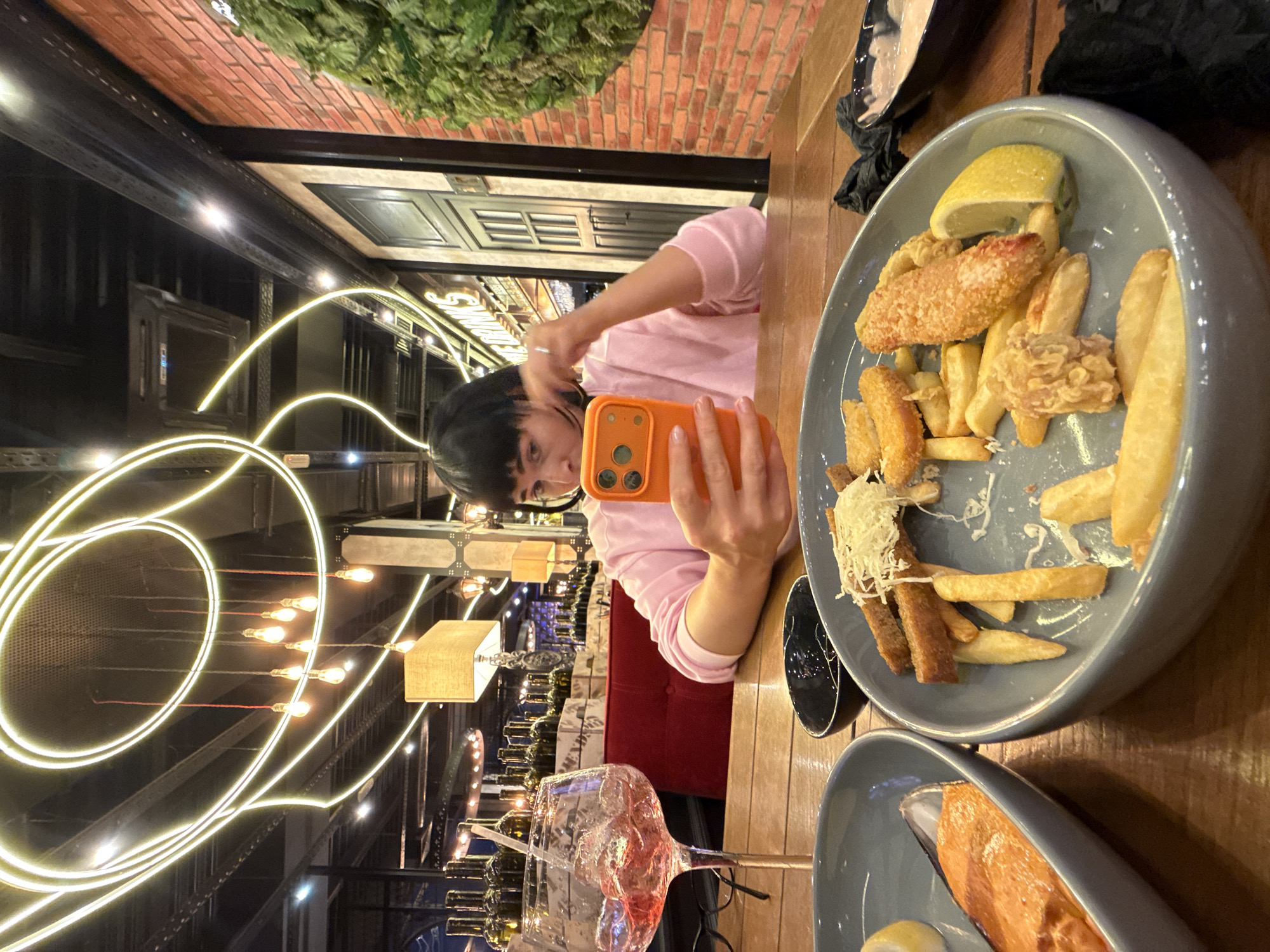 In an interior dining environment, a person is seated at a wooden table, holding an orange smartphone and looking at its screen. The person wears a pink sweatshirt. In front of the person are two plates of food, a glass of pink beverage with ice, and a small black bowl. One plate contains what appears to be fish and chips, with french fries, breaded fish pieces, and a lemon wedge. The other plate holds a piece of salmon and a mussel shell. The background features a brick wall, a green vertical garden, and decorative lighting fixtures overhead. Bottles are visible on shelves in the background.