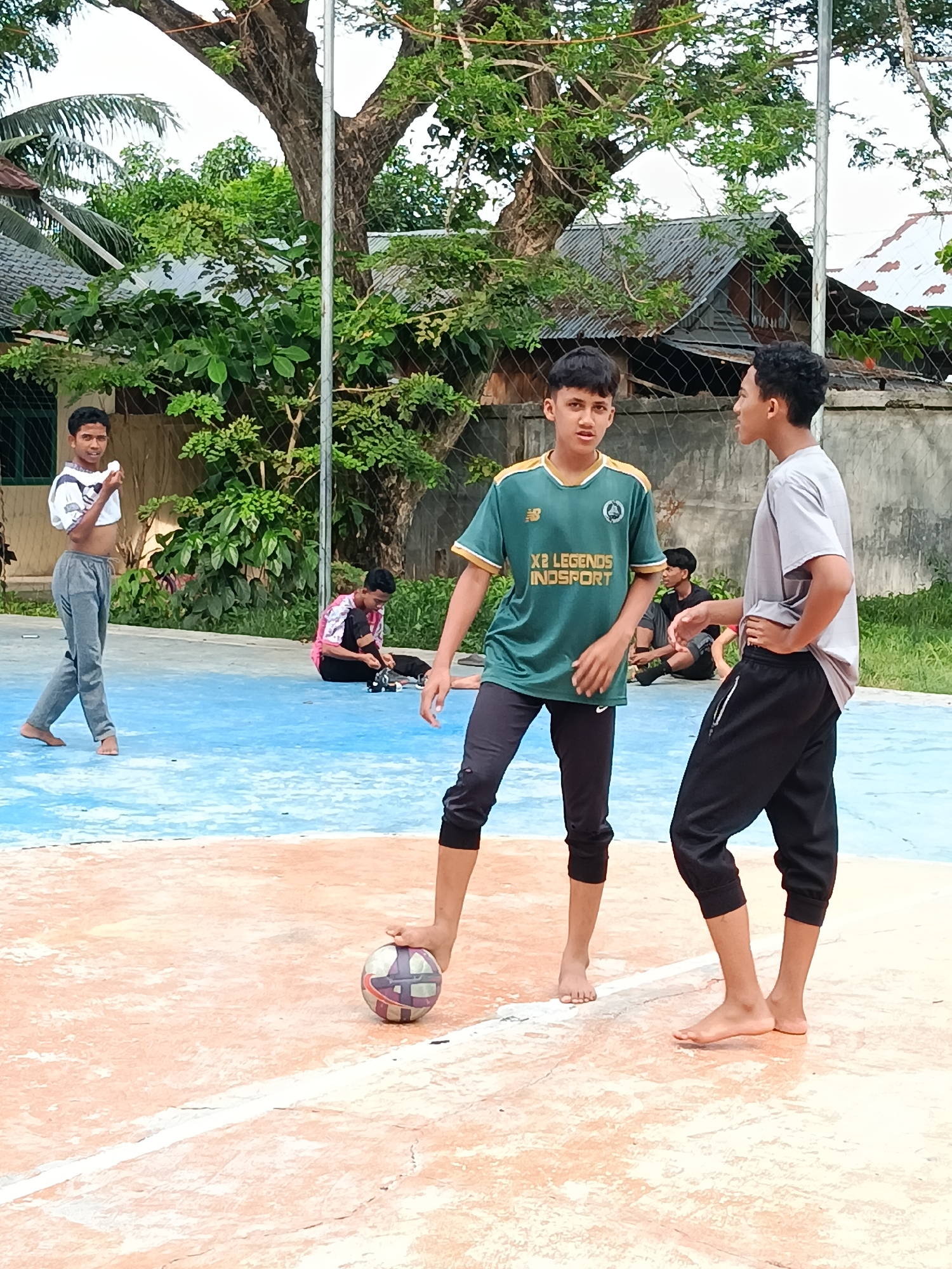 In Lhokseumawe, Indonesia, the scene is a blue and orange outdoor court. A group of young barefoot men are present. One stands with his foot on a soccer ball, facing forward. Another in a gray shirt stands to his right, looking at him. A third man in a white t-shirt is walking away in the background. Two more men sit on the court behind the central figures. Trees and buildings are visible in the background.