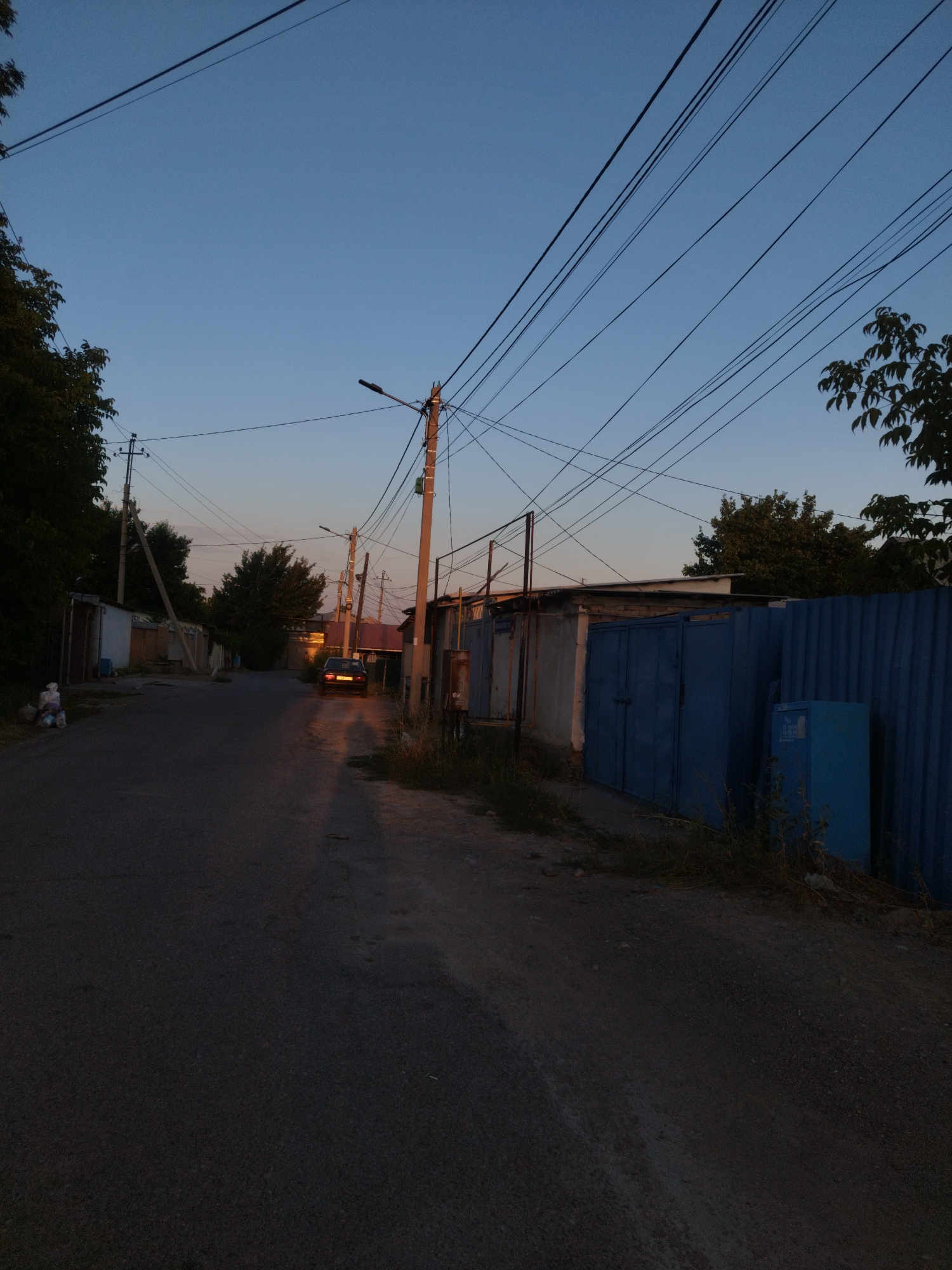 The image depicts a narrow asphalt road in what appears to be a residential or industrial area. The scene is illuminated by the soft, fading light of dusk or dawn, casting long shadows. On the left side of the road, there are several garages with metal doors and some trees. A white trash receptacle is visible near the garages. On the right side of the road, a long, corrugated blue metal fence runs along the property line, with more garages behind it. A blue metal cabinet stands near the fence. A dark-colored car is parked on the road in the middle ground, its brake lights illuminated. Power lines crisscross the sky, supported by wooden utility poles. The sky is a gradient of deep blue to lighter shades towards the horizon, with a hint of orange light from the setting or rising sun. The overall impression is one of a quiet, functional urban environment. The presence of multiple garages and overhead utility lines suggests a developed area. While the exact location within Shymkent, Kazakhstan, cannot be precisely identified from the image alone, the architectural style of the garages and the general streetscape are consistent with such a setting. There are no people clearly visible or engaged in any activity.