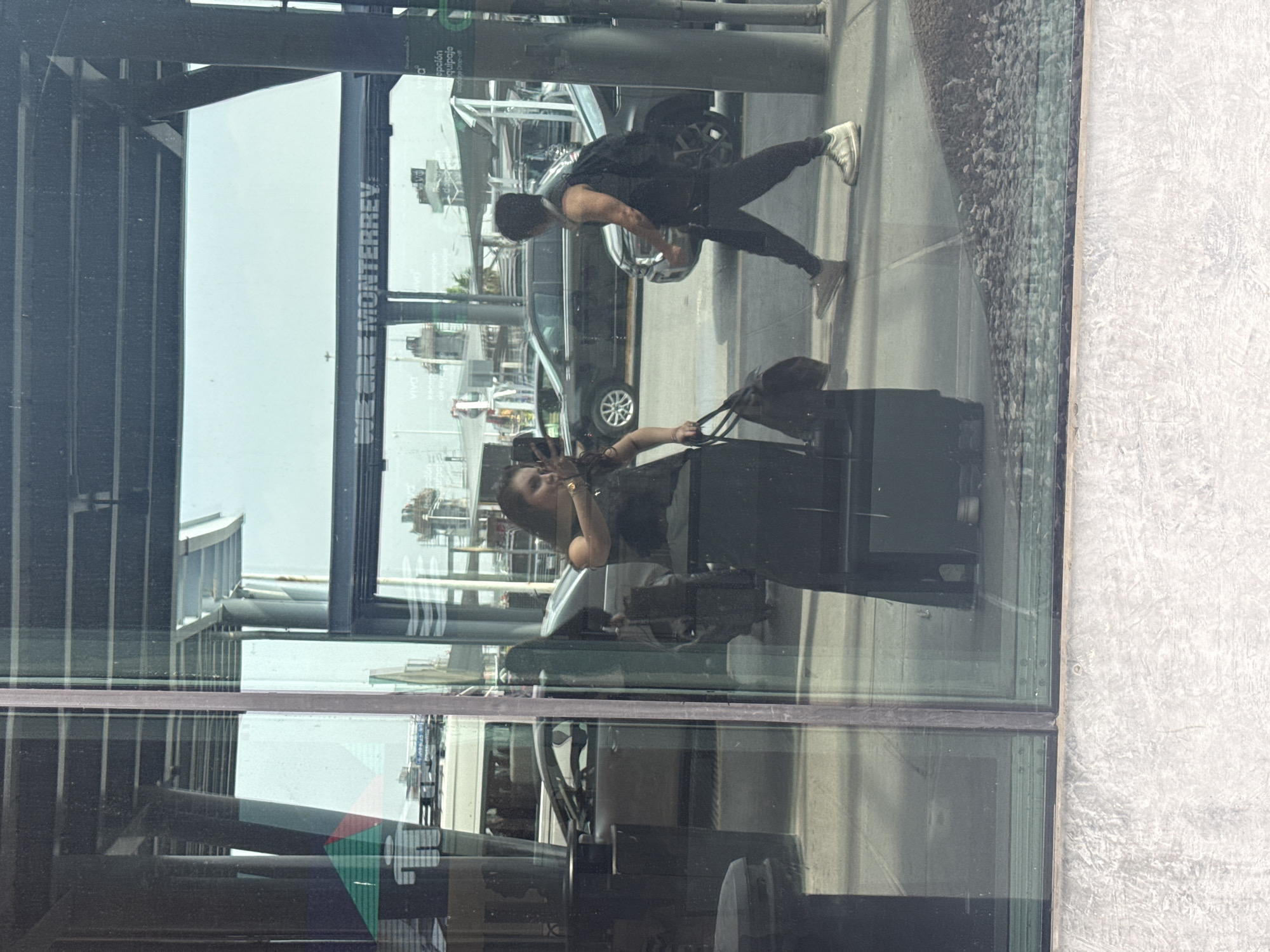 The image captures a bustling scene through a large, reflective glass window, set during a bright, sunny daytime. The overall setting appears to be a modern building, possibly an airport terminal or a similar public transit hub.
In the foreground, a woman with dark hair, wearing a dark top and watch, is visible. She holds a phone to her ear or is taking a picture, and stands beside a black rolling suitcase and a dark handbag. Her presence and luggage suggest she is a traveler waiting or passing through. Her figure appears to be inside the building, reflected and viewed through the glass. To the right, a textured, light-grey concrete wall forms part of the interior or adjacent exterior.
Beyond the glass, an outdoor area is visible. A man, wearing a dark t-shirt, dark pants, and sneakers, is walking or standing. He appears to be carrying a bag on his back. Further back, a dark-colored car, possibly a van or SUV, is parked or driving. The ground outside is paved with a textured, rocky surface.
The highly reflective nature of the glass window creates a complex visual, blending reflections of interior structures, such as dark beams and light ceilings, with the outdoor elements.
Prominently visible on a vertical structure outside the building is partially obscured text that reads "DE CIUDAD MONTERREY," clearly indicating a connection to the city of Monterrey. Another reflection shows text like "VIVA" and fragments such as "cepción" and "equipaje," which likely refer to Viva Aerobus, a Mexican airline, and airport services (reception, luggage). Given the provided context of Apodaca, Mexico, this location is strongly suggested to be Monterrey International Airport.