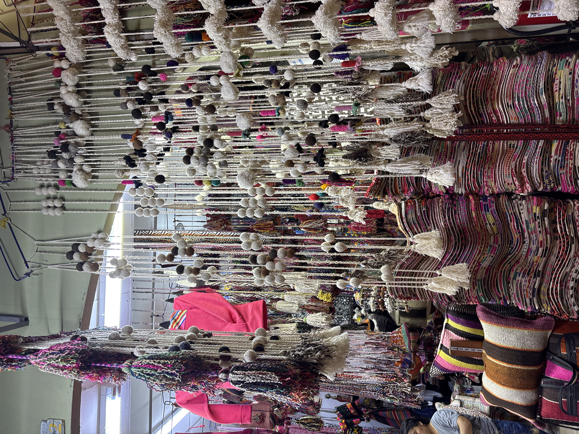 The image depicts a vibrant market stall in Miraflores, Peru, overflowing with colorful textiles and handmade crafts. In the foreground, stacks of intricately woven blankets and pillows with striped patterns in shades of pink, yellow, brown, and white are displayed. Hanging above are numerous strings of yarn adorned with pom-poms and tassels in a variety of colors and textures. A person wearing a grey t-shirt and blue jeans is partially visible on the left side of the frame, facing away from the camera. A bright pink garment, possibly a sweater or jacket, is also visible hanging in the upper portion of the stall. The background is filled with more hanging decorative items, creating a dense and lively visual display of artisanal goods. The scene suggests a bustling marketplace environment where traditional Peruvian crafts are being sold.