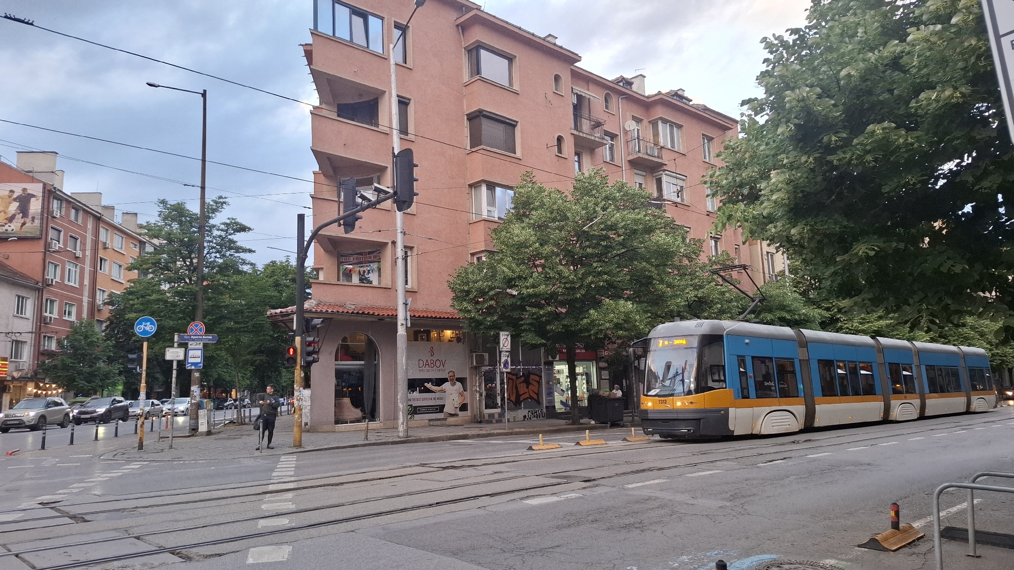 This image depicts an urban street scene in Sofia, Bulgaria, likely at an intersection during an overcast day or late afternoon. A prominent feature is a modern, blue, yellow, and white tram (model "SWING", number "2313") with a destination sign reading "7 И - ЗАПАД" (7 West), positioned on tram tracks on the right side of the frame.
On the right, a distinctive peach-colored building with multiple windows and balconies houses ground-floor businesses. One shop clearly visible is "DABOV SPECIALTY COFFEE," displaying a large cutout of a man holding a coffee cup and the partial slogan "BRING THE BEST COFFEES IN THE WORLD." Above this, another sign partly reads "THE BRITISH HOL" (likely "House" or "Hotel"). A pedestrian in dark clothing is standing near the corner, close to a lamppost and a traffic light showing red.
To the left, a street with several parked and waiting cars runs parallel to the tram tracks. Another multi-story building, lighter in color, is visible further down the street. Green trees are scattered throughout the scene, adding natural elements to the urban environment.
Visible street furniture includes lampposts, traffic lights, and street signs. One sign clearly indicates "Бул. Христо-Ботев" (Bul. Hristo Botev), confirming the location context. Another blue circular sign denotes a bicycle path. The sky is largely cloudy, contributing to the soft, diffused light across the scene.