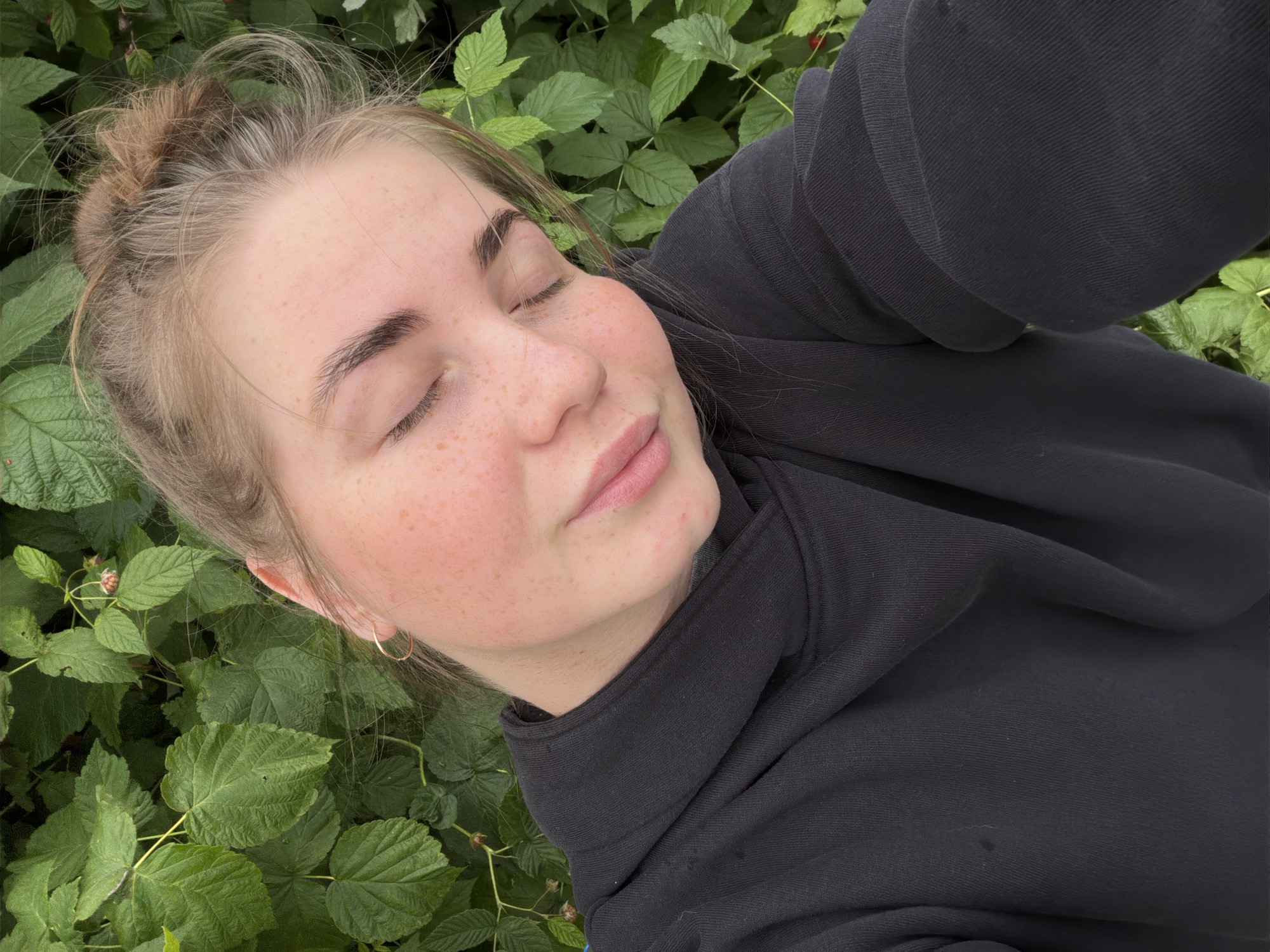 A young woman with blonde hair pulled up in a bun and closed eyes is seen from the chest up, leaning her head slightly to the left. She is wearing a black hoodie and has freckles scattered across her cheeks and nose. Her expression is serene. The background is a dense thicket of green leaves, suggesting an outdoor natural setting. The image does not provide any specific details about the location within the city of Уральское, Russia.