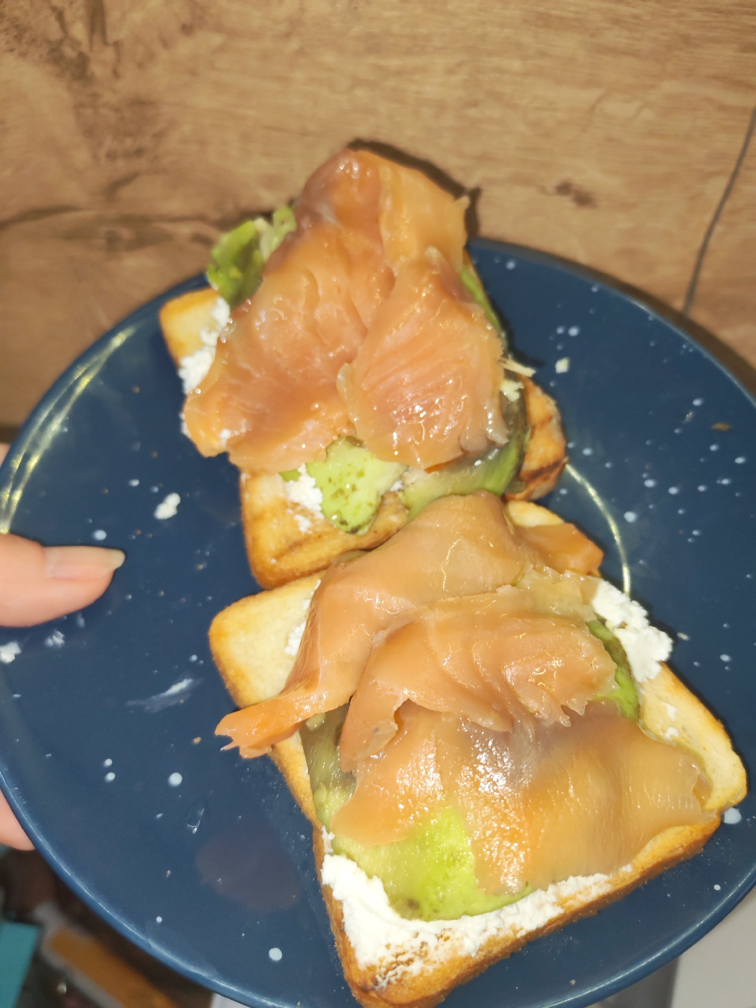 The image presents a close-up view of two open-faced sandwiches, or toasts, resting on a dark blue plate with scattered white speckles. A human hand, with manicured fingernails, is visible on the left side of the frame, holding the plate.
Each toast features a rectangular piece of golden-brown toasted bread as its base. Spread generously over the bread is a white, creamy substance, likely cream cheese or ricotta. On top of this white layer are several slices of vibrant green avocado. The topmost layer consists of multiple thin, glossy slices of smoked salmon, displaying its characteristic orange-pink color, draped over the avocado and cream cheese.
The background of the image is a blurred, warm-toned wooden surface, suggesting a table or countertop. The lighting appears to be indoor, possibly artificial. The overall composition highlights the food, which appears freshly prepared and appealing. No text is visible in the image. This photograph was taken in Izhevsk, Russia.
