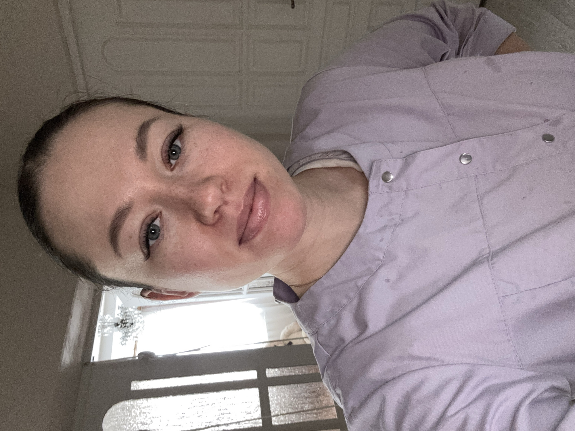 A selfie shot shows a woman with light skin, blue eyes, and dark hair pulled back. She is wearing a lavender-colored garment with visible seams and metal buttons. The background reveals a room interior with a textured ceiling. A window or door with a decorative glass panel is visible, suggesting a domestic or private space. The image location is Ферма, Russia.
