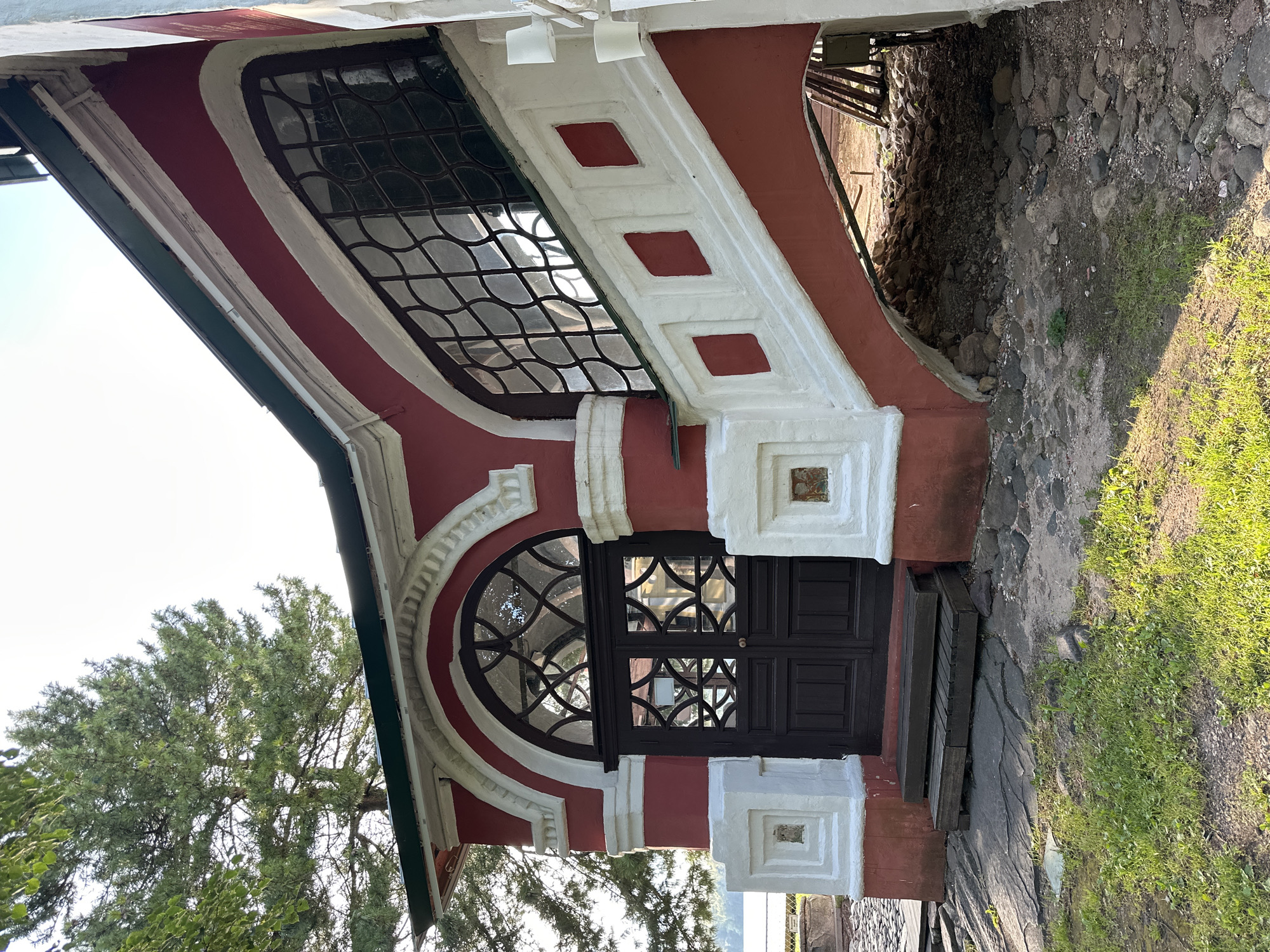 This image shows the exterior of a red and white building with a dark wooden door and an arched window featuring intricate glasswork. To the right, a red staircase with white decorative elements ascends alongside the building. The building is situated in Terpilovo, Russia, with a body of water visible in the background. A large pine tree with green needles is prominent on the left side of the frame, partially obscuring the sky. The ground in front of the building is a mix of grass, stone, and gravel, and there is an arched opening beneath the staircase revealing a rocky, debris-filled area.