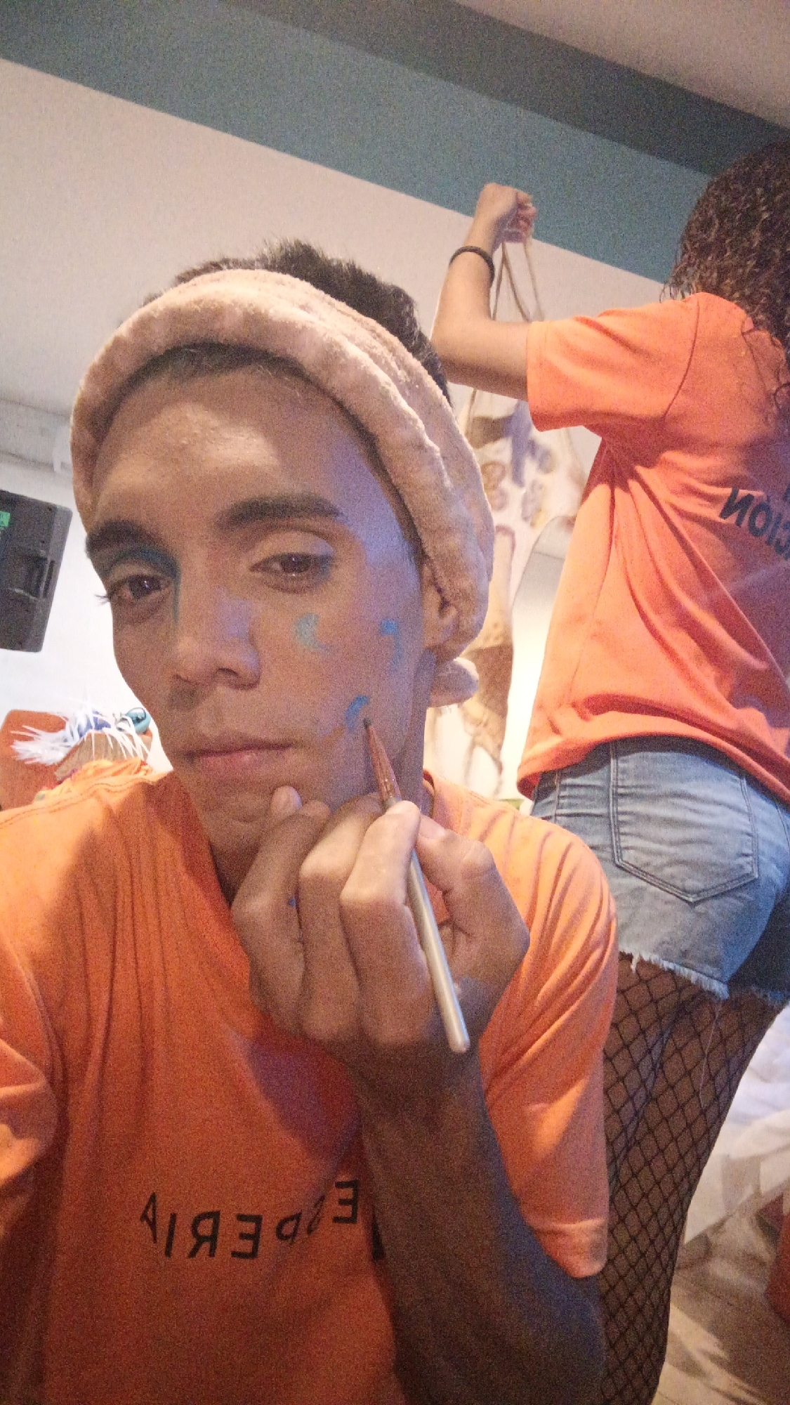 A person is shown applying blue paint to their face, with a peach-colored headband visible. The person is wearing an orange t-shirt with black text printed backward, reading "EXPERI," the remainder of the word obscured. In the background, another person, also wearing an orange shirt and denim shorts over black fishnet stockings, appears to be holding a bag. The location appears to be indoors. Given the image context, the location is most likely Playa El Agua, Venezuela.