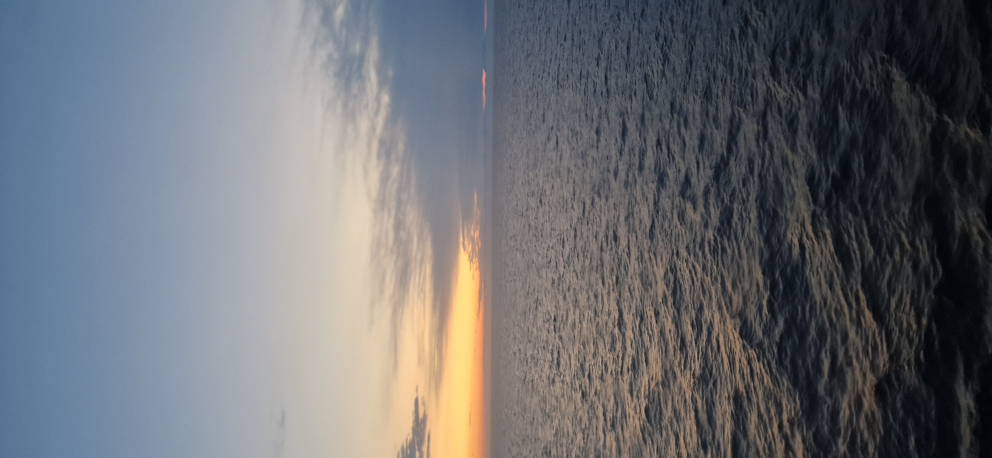 The image depicts a serene seascape at dusk. The sky transitions from a clear, pale blue at the top to hues of orange and yellow near the horizon, where scattered clouds are visible. The ocean occupies the lower half of the frame, with gentle waves reflecting the fading light. The overall impression is one of tranquility and vastness. The location is described as Unknown, Croatia, but no specific landmarks or urban elements are visible to confirm this.