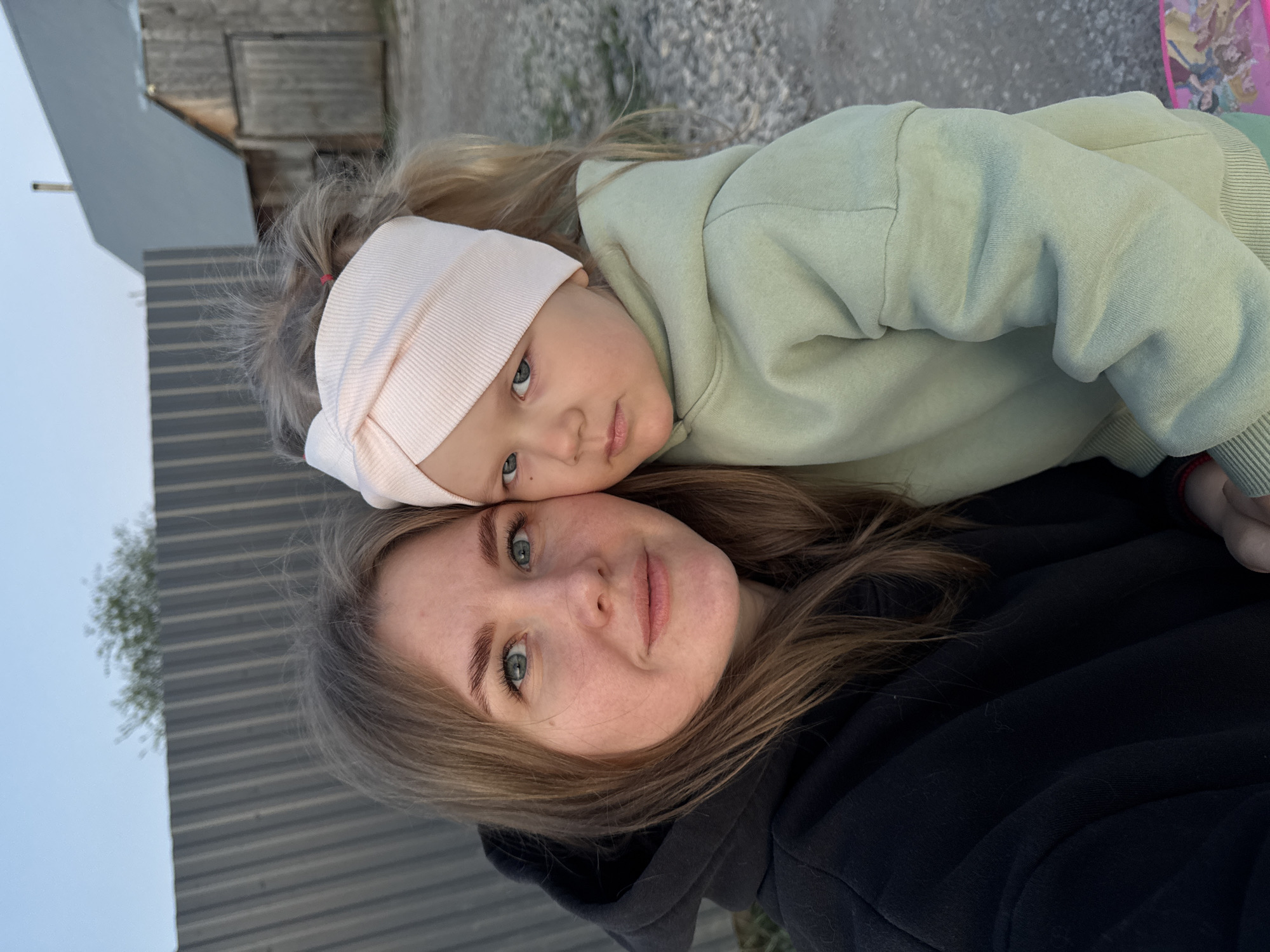 A woman and a young child, possibly a mother and daughter, are posing for a selfie outdoors in Горный, Russia. The woman, with light brown hair, is wearing a black hooded sweatshirt. The child, resting her head on the woman's forehead, has blonde hair and is wearing a light green hooded sweatshirt and a light pink headband. The background includes a corrugated metal fence, a building, and a gravel surface. The sky is visible in the upper left corner.