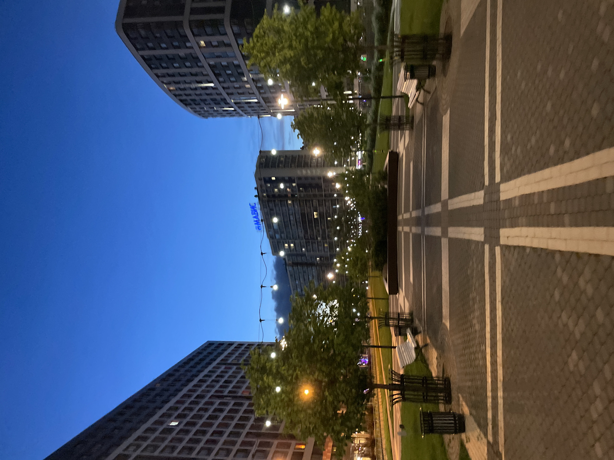 This image depicts an urban park scene at twilight in an unknown location in Russia. In the foreground, a paved walkway with white striping leads towards a central grassy area dotted with young trees in planters. Several park benches and trash receptacles are strategically placed along the walkway and grassy areas. String lights are visible overhead, casting a soft glow across the scene, and streetlights illuminate the pathways.
Dominating the background are two large, modern apartment buildings. The building on the left is a monolithic structure with a grid of windows. The building on the right has a curved facade, also featuring numerous windows. A prominent blue sign spelling "АМАРВИС" in Cyrillic is visible on the facade of a building in the middle distance. The sky is a deep twilight blue, with some faint clouds visible. The overall atmosphere is serene and tranquil, suggesting an evening setting in a well-maintained urban development. No people are visible in the image.
