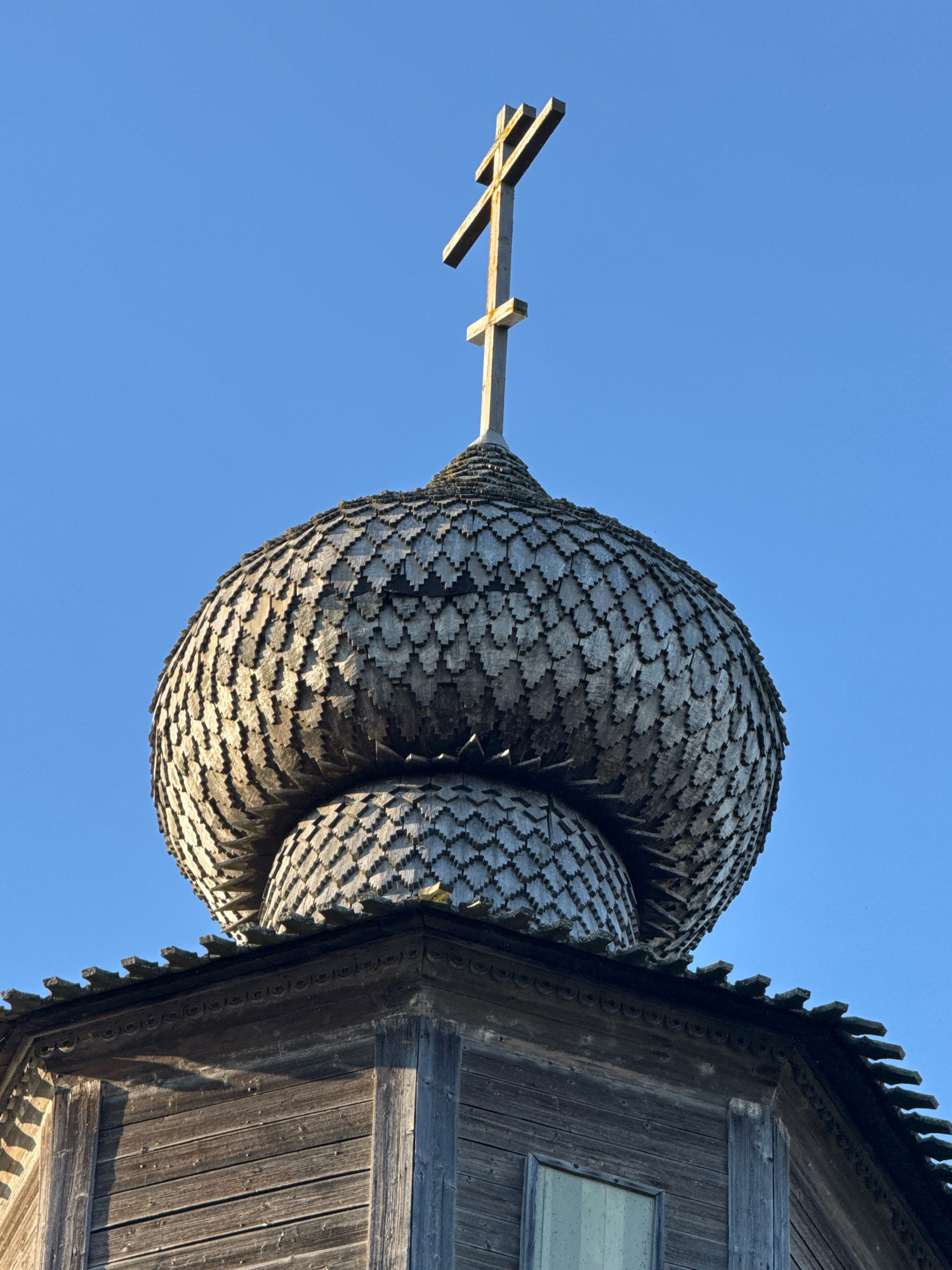 The image shows the roof of a wooden Orthodox church in Torzhok, Russia. The roof is topped with a traditional Orthodox cross. The domes of the church are covered with wooden shingles that resemble scales. The wooden walls of the church are visible below the domes. The sky is clear and blue.