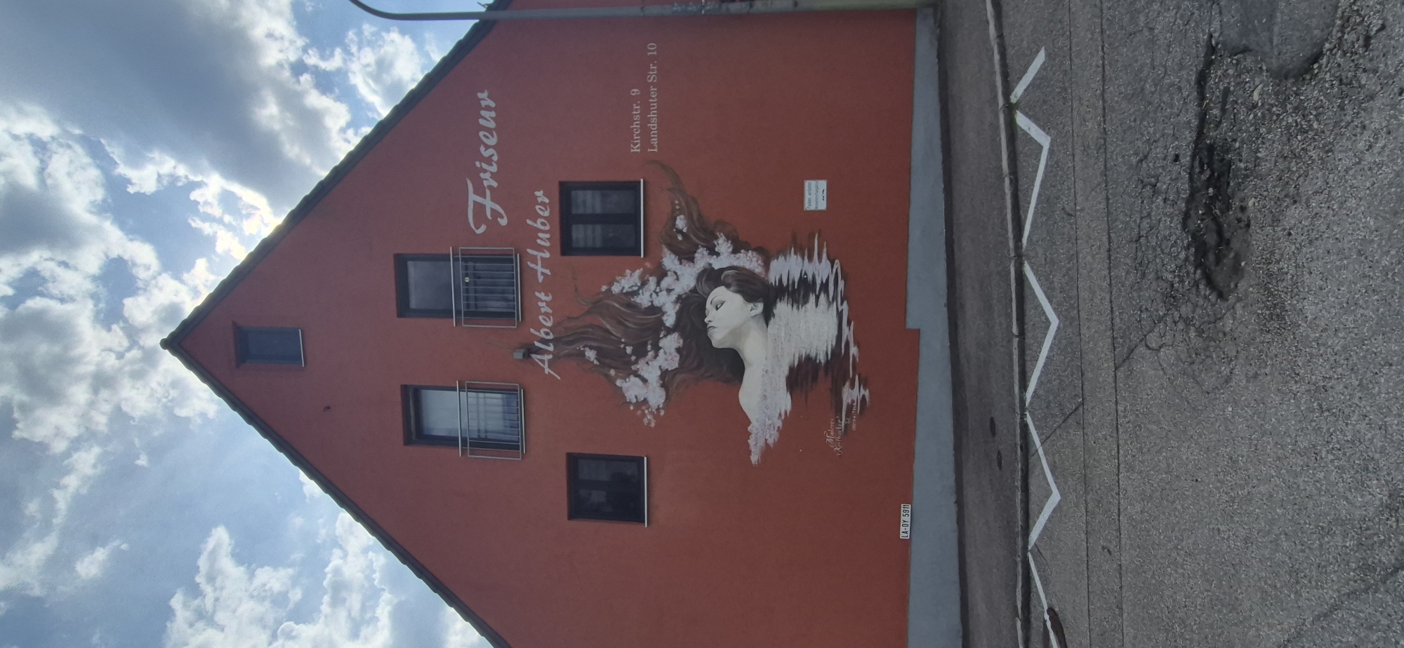 The image depicts the facade of a building painted with a mural. The mural features a woman's face and upper body emerging from water, with her hair flowing outwards and adorned with white flowers. Above the mural, the words "Friseur Albert Huber" are painted in white script. To the right of the mural, an address is listed: "Kirchstr. 9 Landshuter Str. 10". A small white sign with text is also visible on the wall to the right of the mural. The building is painted a vibrant red and has several windows and balconies. The sky above is blue with scattered white clouds. In the foreground, a street with white zig-zag markings and a pothole is visible. The building is located in the city of Velden, Germany.