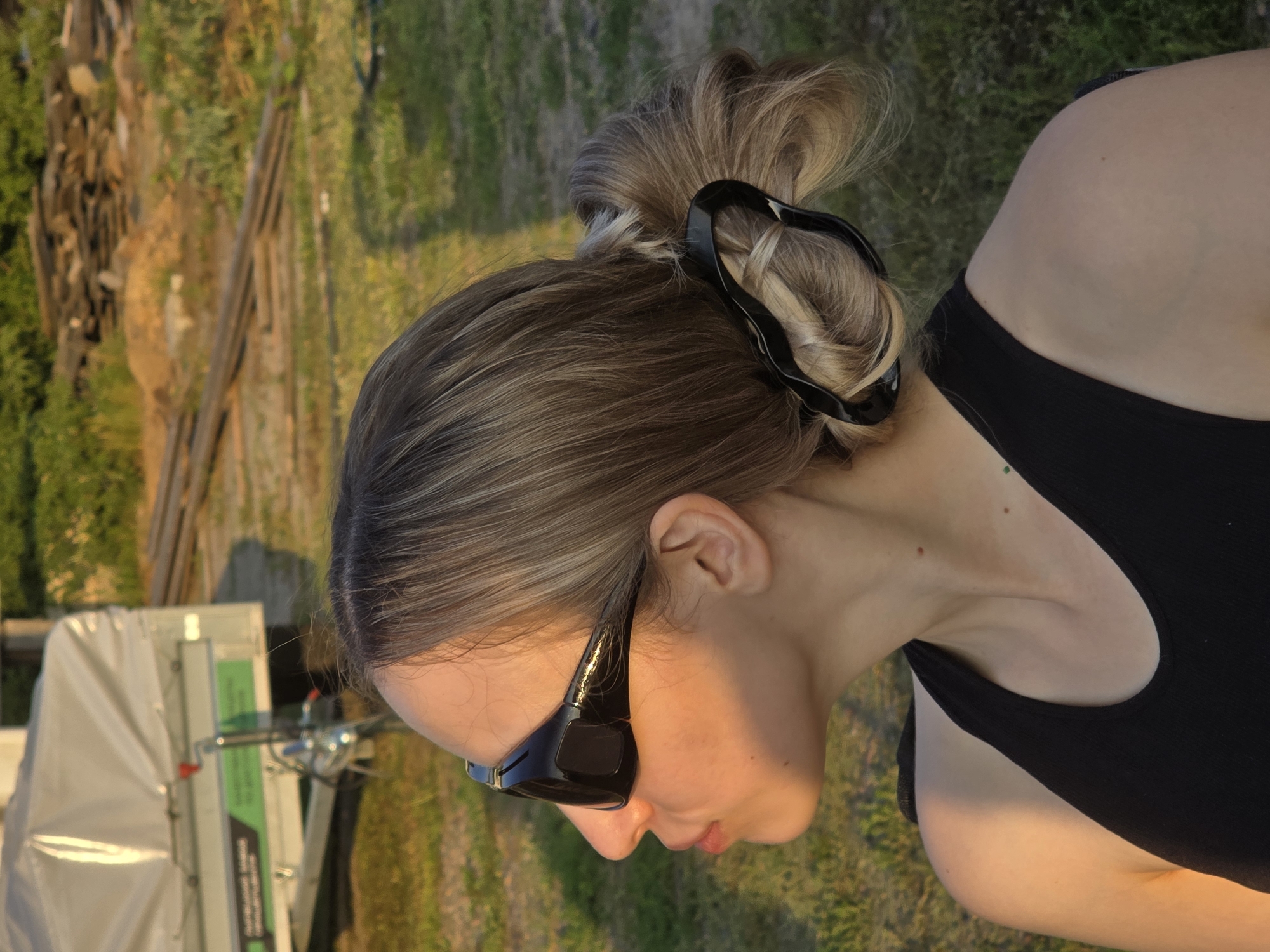A young woman with blonde hair pulled back in a bun with a black claw clip wears large black sunglasses and a black tank top. She is outdoors, with a trailer in the background. The sun casts a warm glow on her face and shoulder. The environment is grassy with some wooden debris in the background.
