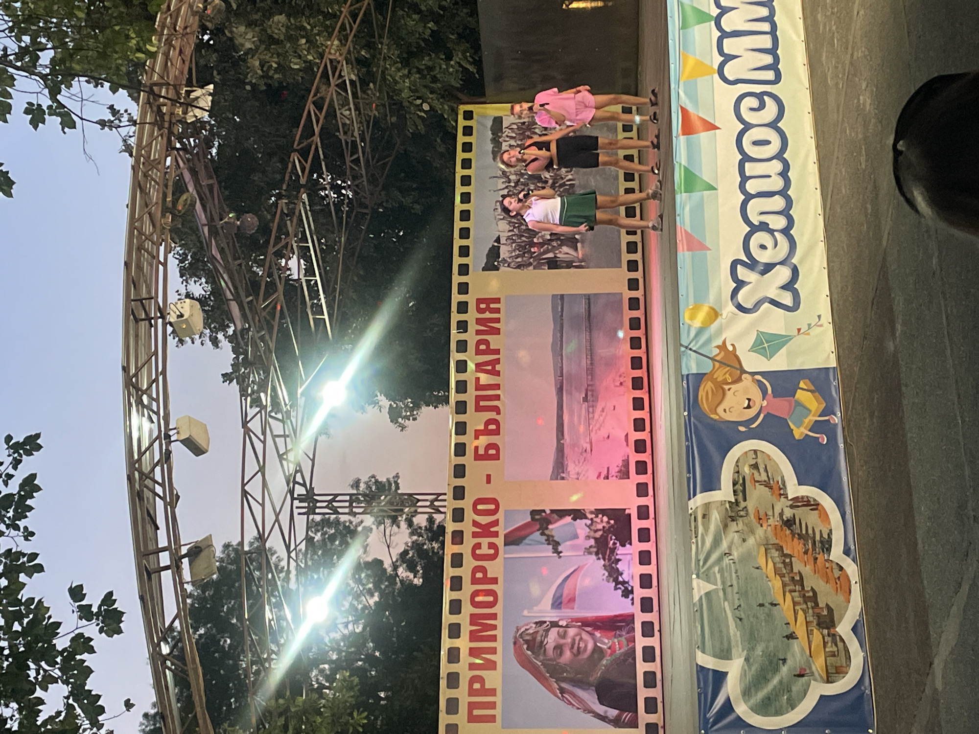 The image shows an outdoor stage in Kiten, Bulgaria, likely during an evening event. In the foreground, three young girls are singing into microphones on a stage, dressed in casual attire. Behind them, a large banner displays images of a woman in traditional dress, a beach scene, and a crowd of people. Below this banner, another banner features a cartoon child on a swing and colorful pennants. The stage structure is visible, with lights and scaffolding against a twilight sky and silhouetted trees. The overall scene suggests a public performance or festival.