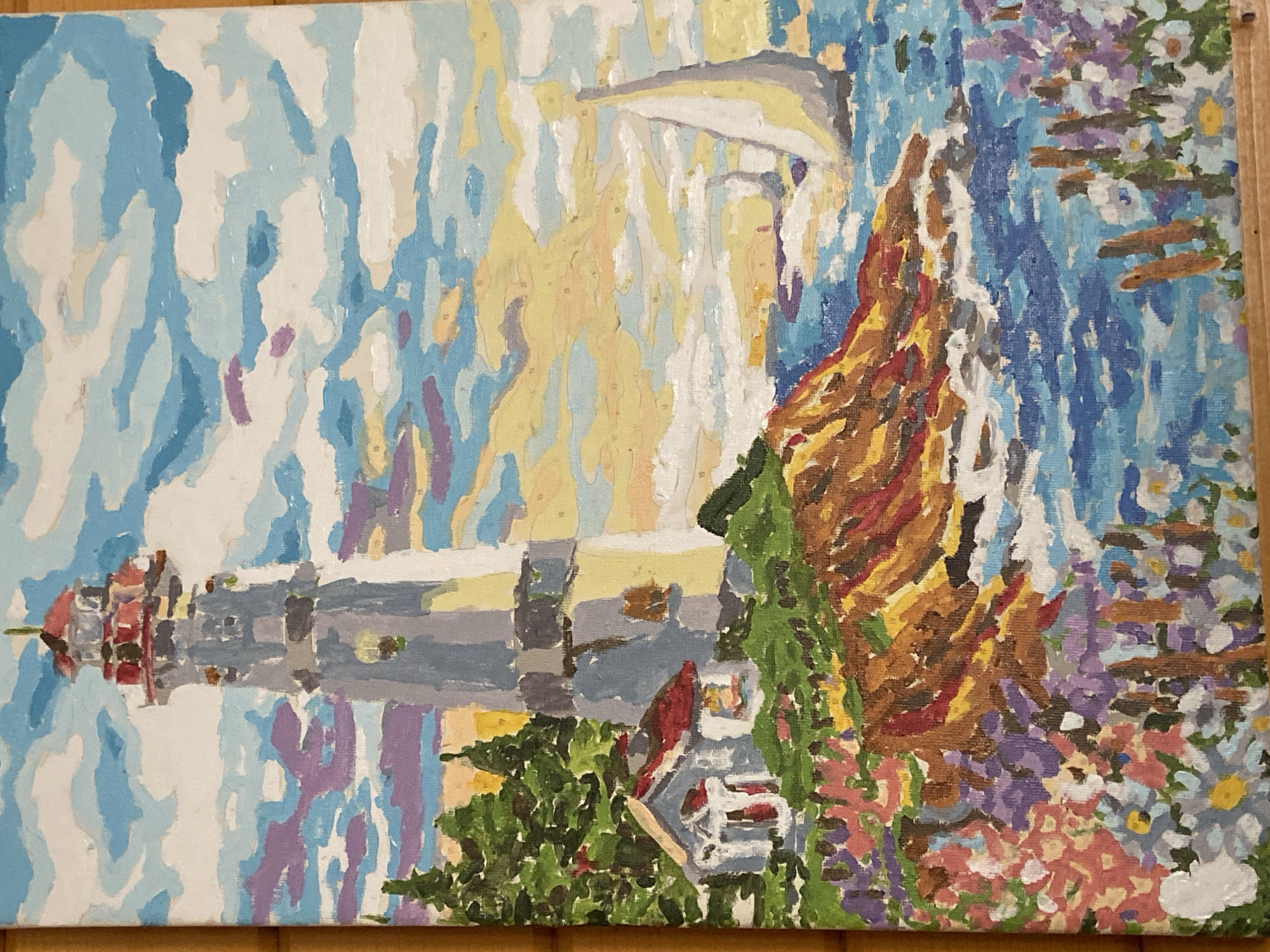 The image depicts a paint-by-numbers style painting of a lighthouse scene. The lighthouse is positioned on the left side of the painting, with a house situated at its base. The landscape features a rocky cliff to the right, adorned with a variety of plants and flowers in shades of green, purple, and pink. The sky is rendered in an impressionistic style with various shades of blue and white, while a section of the landscape is washed with yellow hues, likely representing sunlight. The painting exhibits a deliberate, sectioned approach, evident from the distinct blocks of color that form the overall image.