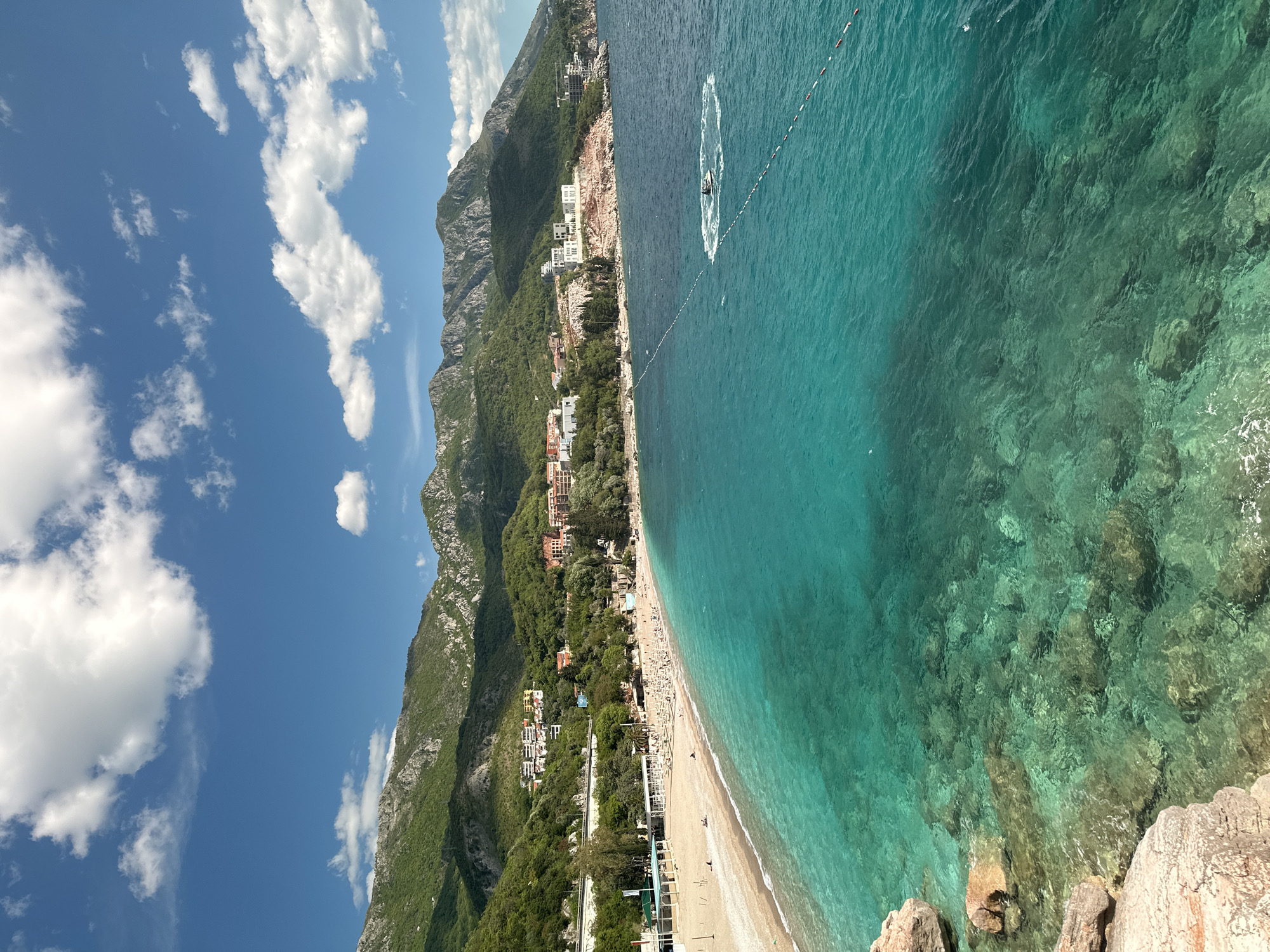 This image presents a stunning aerial view of a coastal landscape at Kamenovo, Montenegro, on a clear and sunny day. The scene is dominated by natural beauty, featuring a long beach, an incredibly clear sea, and dramatic mountains under a bright blue sky with scattered white clouds.
In the background, steep, verdant mountains rise, covered in dense green vegetation and showcasing rugged rocky sections. Along the base of these mountains, a stretch of development is visible, with numerous buildings, some appearing to be hotels or residences, nestled amidst trees and foliage.
The central feature is a wide, light-colored beach, likely a mix of sand and pebbles. It is dotted with many people relaxing, sunbathing, and enjoying the pleasant weather, along with scattered beach chairs and umbrellas.
Adjacent to the beach, the crystal-clear sea stretches out, exhibiting vibrant shades of turquoise and clear blue. The transparency of the water allows the rocky seabed to be clearly visible, especially in shallower areas. A line of red and white buoys extends across a section of the water, likely demarcating a swimming zone. Further out, a small watercraft, possibly a jet ski, is actively moving, leaving a distinct circular white wake on the water's surface, indicating recreational activity.
The foreground shows rocky outcrops, suggesting the photograph was taken from an elevated viewpoint, such as a cliff or hillside, overlooking the bay. The strong daylight and lack of shadows indicate midday or early afternoon. No discernible text is visible within the image.