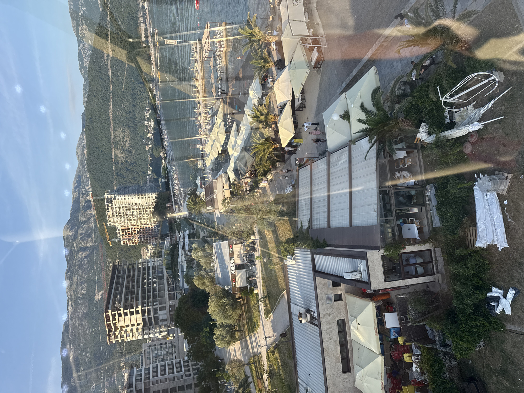This media file presents a panoramic view of a vibrant coastal town, identified as Budva, Montenegro, likely taken from an elevated perspective through a window, given the visible reflections. The scene is set under a clear blue sky, with a long range of verdant mountains dominating the background on the left.
The bustling coastline unfolds from left to right, featuring a marina filled with numerous boats and yachts. Adjacent to the marina is a lively beach area, densely covered with rows of white sun umbrellas and sun loungers. Several people are visible relaxing on the beach, swimming in the calm waters, or strolling along the shore.
Running parallel to the beach is a pedestrian promenade and a road, flanked by a variety of buildings. Many of these structures house restaurants and cafes, indicated by extensive outdoor seating shaded by white umbrellas. Palm trees are interspersed throughout the area, enhancing the resort atmosphere. Text visible on some umbrellas includes "Coca-Cola" logos and partial words like "KRA..." and "RES...". A sign near the water reads "ASAS". Further inland, a building displays "PIZZA" and "COCO".
The landscape also shows areas of ongoing development, with several multi-story buildings under construction, suggesting growth in the region. In the immediate foreground, a grassy area contains stored or discarded outdoor furniture and equipment, including a white, arch-shaped structure.
The overall impression is of a sunny and active day in a popular Mediterranean resort. The bright sunlight casts distinct shadows, indicating late morning or early afternoon. People are engaged in various leisure activities, from dining outdoors to enjoying the beach, contributing to a lively and picturesque coastal scene.
