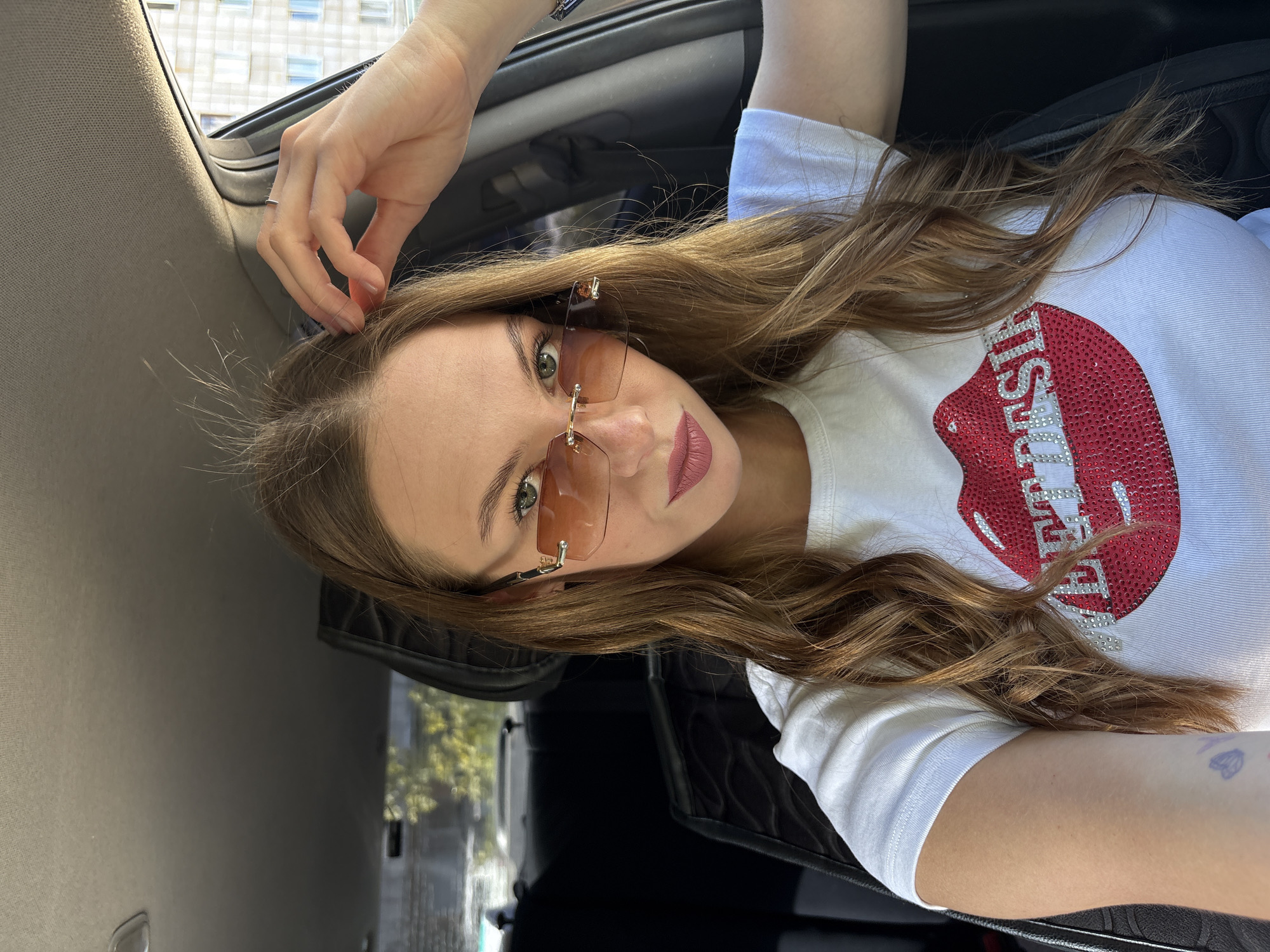 A young woman with long, wavy brown hair is seated in the driver's seat of a car. She is wearing sunglasses with amber lenses and a white t-shirt adorned with a sequined graphic of red lips and the word "SWEET DESIRE" in rhinestones. Her right hand is raised, touching her hair. The interior of the car is visible, including the car seat headrest and part of the dashboard. Outside the car, a building with multiple windows can be seen. The image appears to have been taken during daylight.