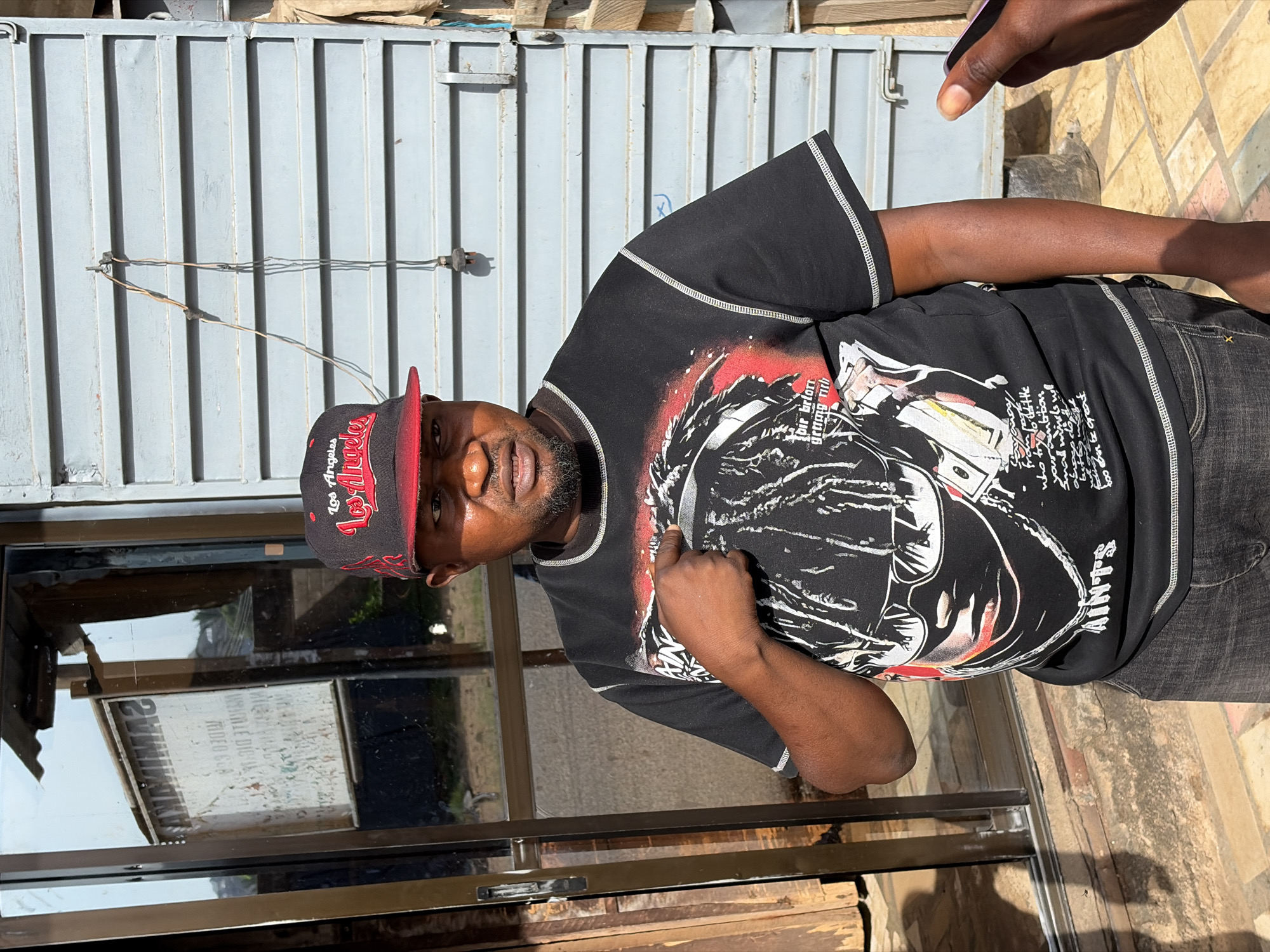 A man wearing a black "Los Angeles" baseball cap and a black t-shirt with a complex graphic print stands in front of a glass storefront with a metal frame. The store's sign is visible through the glass, though the text is illegible. The man points towards himself. Behind him, a corrugated metal wall is visible. The ground appears to be made of stone or weathered concrete. A person's hand is visible in the upper right corner. The location is presumed to be Jos, Nigeria.