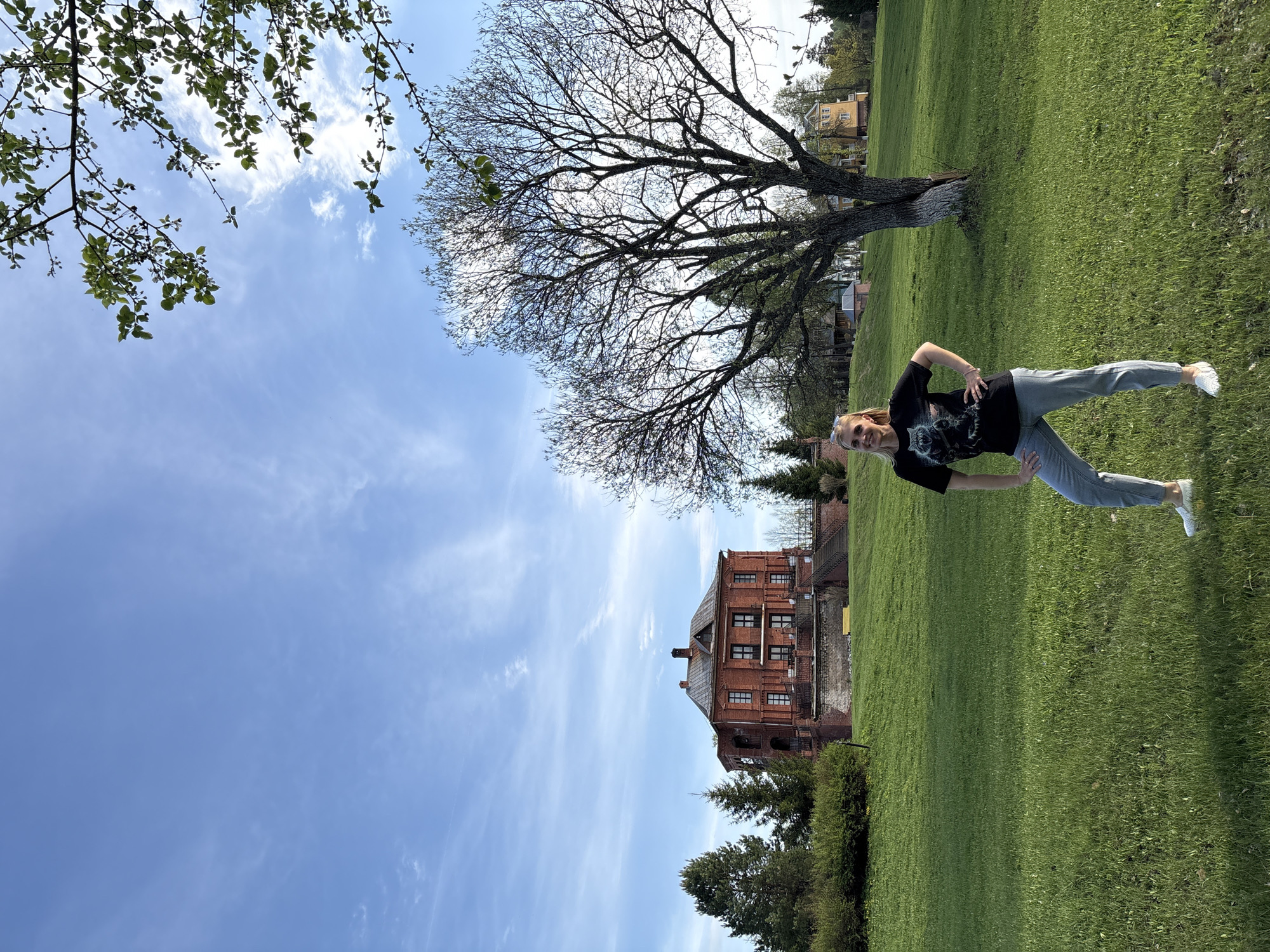 The image shows a woman in athletic wear posing on a grassy lawn. She wears a black t-shirt with a wolf graphic, grey sweatpants, and white shoes. She has short blonde hair. Behind her is a large tree, and in the background is a red brick building. The sky is blue with scattered clouds. This scene appears to be taken in Zavod Mikhailovskii, Russia.