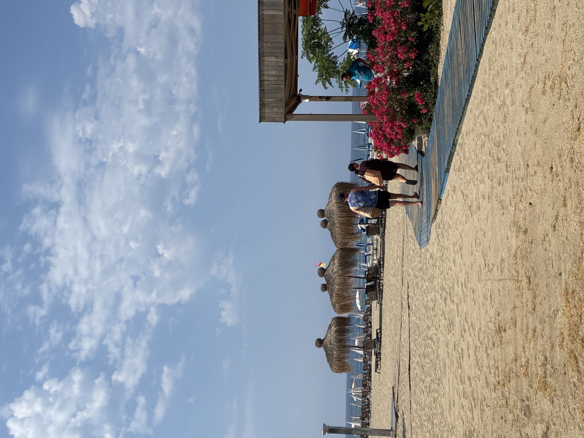 The image depicts a sandy beach scene in Alanya, Turkey, under a clear blue sky with scattered white clouds. In the foreground, a blue wooden boardwalk leads towards the right side of the frame. Two individuals, a man and a woman, are walking away from the viewer on this boardwalk. The man wears a blue and white patterned tank top and black shorts, carrying a straw bag. The woman is dressed in a dark dress and carries a brown shopping bag.
To the right, a wooden structure, likely part of a beach bar or cafe, is visible, with a man in a blue t-shirt standing near it. In front of this structure is a vibrant display of pink flowering bushes. Behind the boardwalk and the individuals, the beach is lined with rows of empty sun loungers and straw umbrellas, suggesting a peaceful, off-peak moment. Further back, the turquoise waters of the sea are visible, with several more umbrellas dotting the coastline and a few small boats in the distance. The overall atmosphere is serene and summery, characteristic of a Mediterranean beach resort.