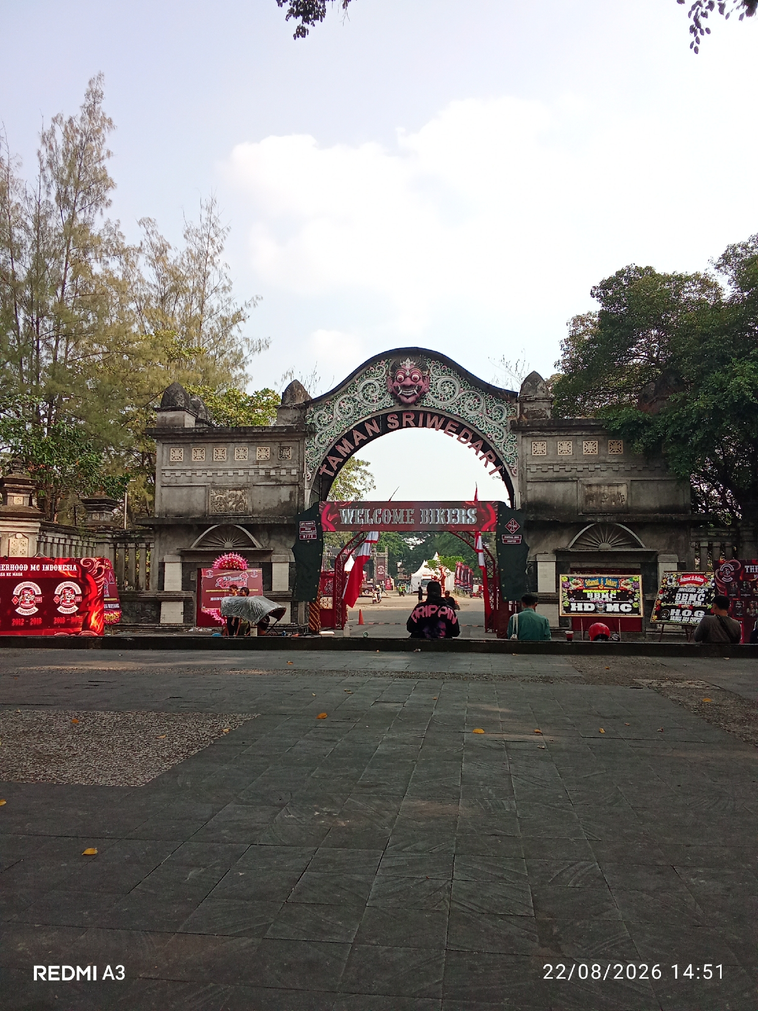 The image shows the entrance gate of Taman Sriwedari in Surakarta, Indonesia. The gate is adorned with traditional Javanese carvings and a large archway. "WELCOME BIKERS" is prominently displayed on a banner under the arch. Several individuals are seated on the ground in front of the gate, some facing away from the camera and others to the side. Red banners with logos, possibly related to motorcycle clubs, are visible on the left side of the entrance. The ground in front of the gate is paved with dark square tiles, and a few fallen leaves are scattered across it. The sky above is mostly clear with some scattered clouds. A date and time stamp, "22/08/2026 14:51," is visible in the lower right corner of the image, along with "REDMI A3" in the lower left corner.