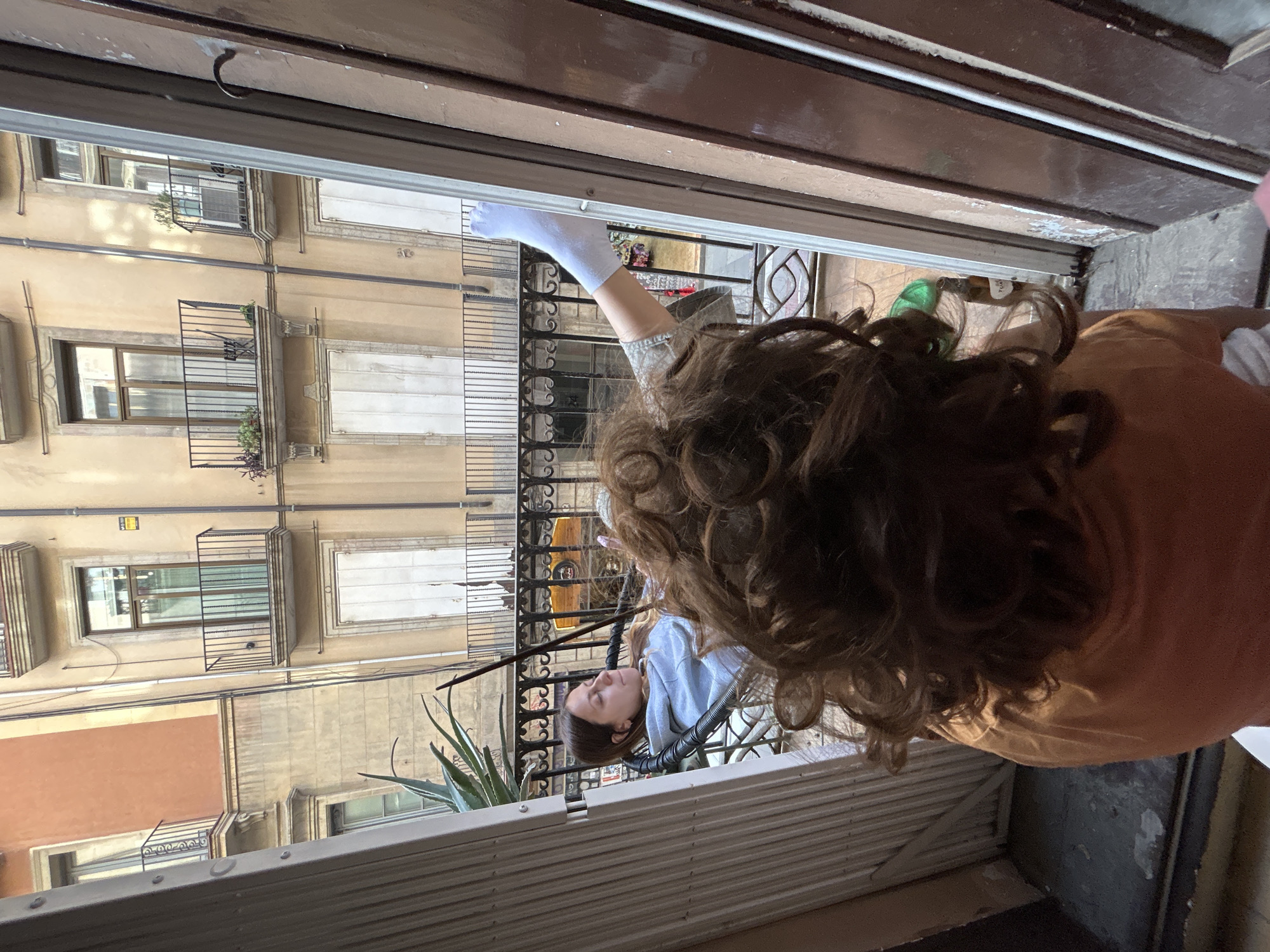 The image, initially rotated 90 degrees clockwise, presents a view looking out from an open doorway or window onto an urban scene, characteristic of Barcelona, Spain.
In the foreground, dark brown door or window frames are visible at the top and left, along with a white frame and part of a white louvered blind or shutter system in the lower section. A person with long, dark, curly hair, wearing an orange/tan top, is partially visible in the bottom right, leaning out. Their back is mostly towards the viewer, and their head is turned towards the left, as if looking down or observing something. A hand, covered in a white glove, extends from their arm, reaching outwards, possibly for cleaning or adjusting something.
Below and to the left, on a lower balcony of a neighboring or the same building, another person is seen resting in a black chair. This individual has brown, relatively straight hair and is wearing a light blue top. Their eyes appear closed or they are looking upwards, conveying a sense of relaxation or sleep. Green, succulent-like plants are visible on this balcony.
The background reveals a dense urban environment with multi-story residential buildings. These structures feature light yellow or beige stucco facades, multiple windows, and black wrought iron balconies, some adorned with potted plants. Utility pipes run horizontally across the building exteriors. The scene suggests a bright, perhaps sunny, daytime. No clearly legible text is visible, though a small, unreadable sign is on a distant building wall.