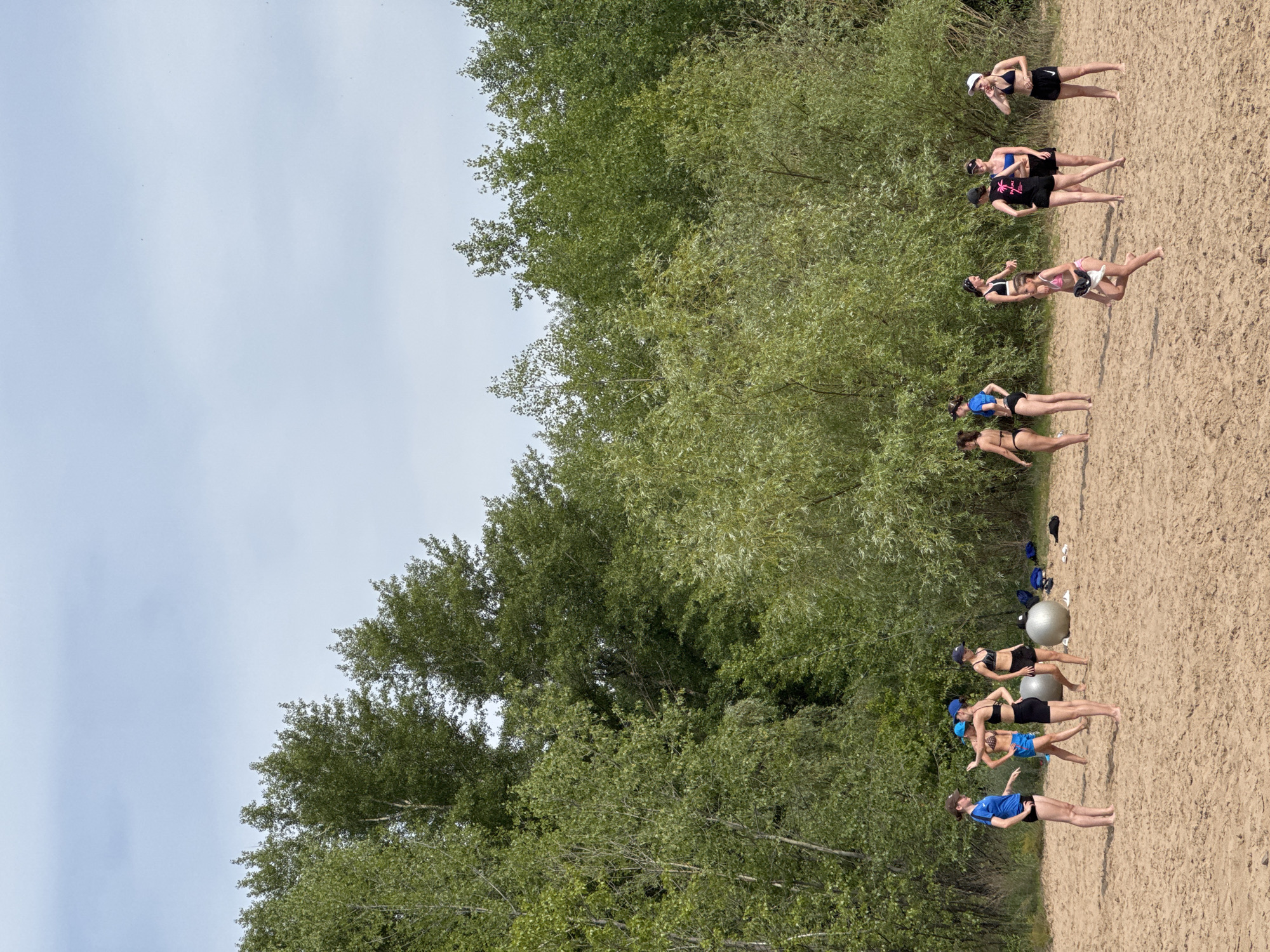A group of women are lined up on a sandy beach, with trees and foliage forming a backdrop. They appear to be engaged in a workout or training session, with some individuals in athletic wear and others in swimwear. One woman in the front wears a blue t-shirt and black shorts, while others behind her are in bikinis or sports bras and shorts. Two large exercise balls are visible, suggesting a fitness activity. The lighting suggests it is daytime, with bright sunlight creating shadows on the sand. The sky is overcast with a light blue hue. The overall atmosphere appears to be one of focused activity, with the women in various stances, some appearing to be in mid-exercise or movement. The setting is clearly outdoors, on a beach with a clear delineation between the sand and the densely vegetated area.