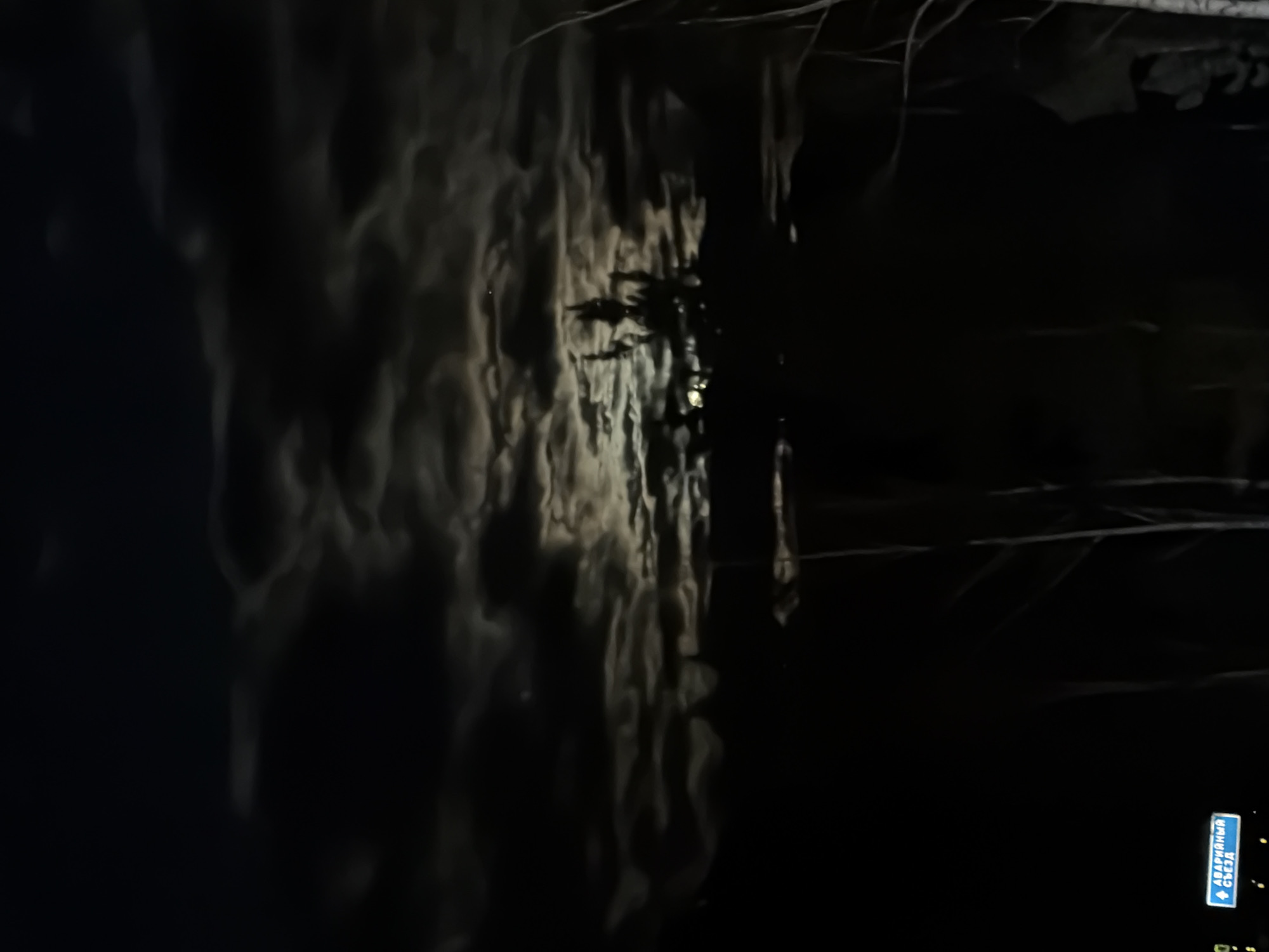 The image depicts a nighttime scene near Слизнево, Russia. There is a street sign in the bottom right corner with directional information. Adjacent to the sign is darkness. To the left of the dark section, a white wall rises with vertical striations along its length. The left side of the wall is brighter, transitioning towards a darker area, while a thin line of vegetation sits atop the wall and along the right-hand side. Bare branches extend diagonally across the top of the frame. The environment is dark, with a lack of illumination.