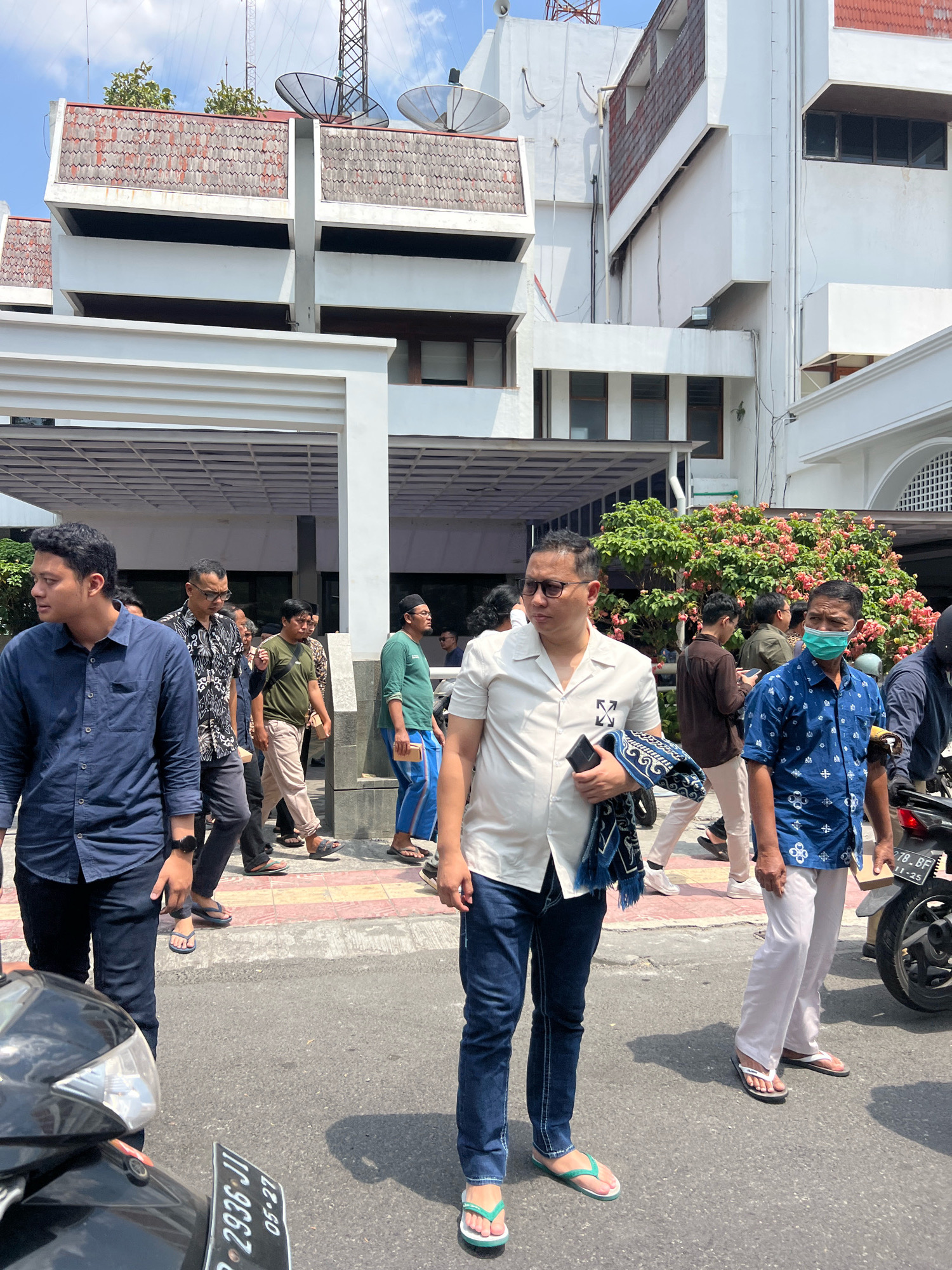 The image shows a group of men walking on a street in front of a white building. The building appears to be a government or institutional building, with satellite dishes visible on the roof. Some of the men are wearing casual clothing, while others are dressed more formally. One man in the foreground is wearing a white shirt and jeans, and he is holding a patterned cloth. Another man in a blue patterned shirt and white pants is wearing a face mask. The scene suggests a public gathering or event. The street is paved, and there are some trees and flowering bushes in the background. The sky is blue with some clouds. There is a motorcycle parked on the right side of the image. The license plate on the motorcycle indicates it is registered in Indonesia. Based on the architecture and the presence of other elements, the location is likely in the city of Yogyakarta, Indonesia.