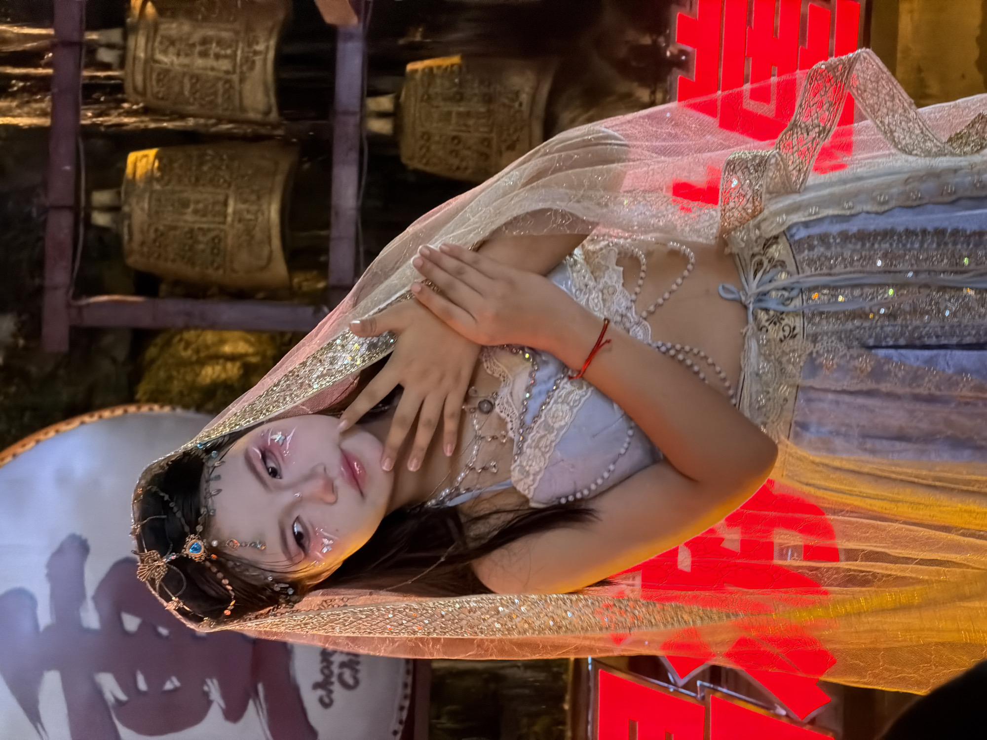 A young woman, adorned with elaborate makeup and a traditional outfit featuring a veil, is captured in a studio or staged setting. The outfit consists of a light blue top with intricate lace detailing, a pearl necklace, and a shimmering, gold-trimmed veil. Her makeup is dramatic, with exaggerated eyeliner, blush, and iridescent accents on her cheeks and eyelids. She wears a delicate headpiece adorned with beads and a blue gem.
The background hints at a cultural or festive event. Two large, ornate golden bells are visible on shelves in the upper left. In the foreground and to the right, large, bright red Chinese characters are illuminated, suggesting a celebratory context, possibly a wedding or festival, given their usual application. The red characters appear to be part of a backdrop or stage decoration. The floor or surface she is lying on is reflective, showing a distorted reflection of her and the surrounding elements. The overall lighting is warm and dramatic, with a focus on the subject. The scene appears to be indoors or a covered outdoor space, possibly at night or in dim lighting conditions, given the artificial illumination. There are no clear indications of weather. The overall mood is one of artistic presentation and cultural symbolism. The visible text in red are Chinese characters, but they are too stylized and large to identify specific meanings without further context. There is also faint text on a circular object in the lower left corner, partially obscured, that appears to read "chan chi".