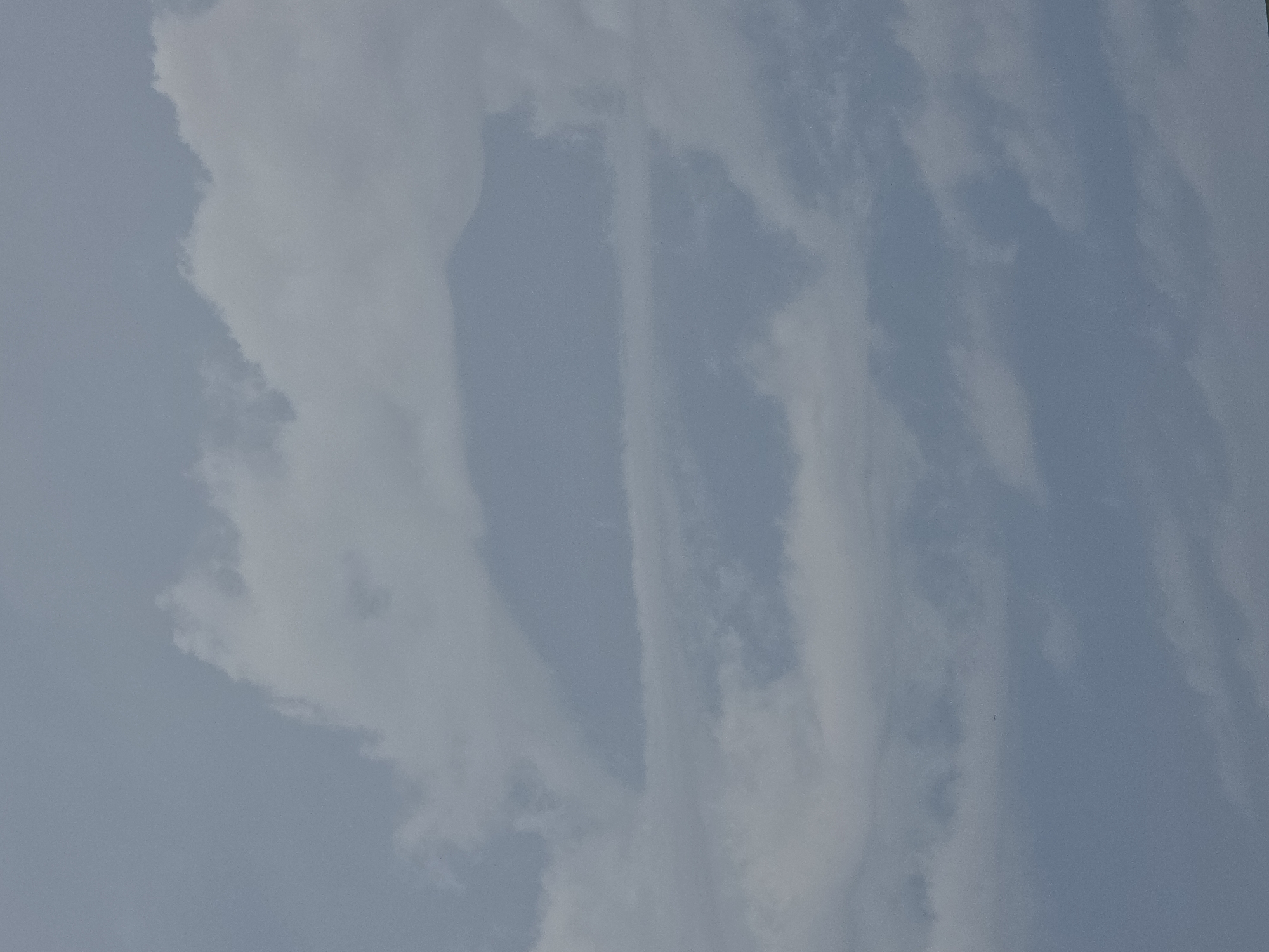 The image shows a cloudy sky above Stanghella, Italy. The clouds are dense and gray, with some lighter patches suggesting sunlight breaking through. In the distance, the outline of a mountain range is faintly visible through the clouds, suggesting a possible elevation in the Stanghella area. There are no people, subjects, or objects clearly discernible within the frame of the image, focusing solely on the atmospheric conditions. The scene is neutral and devoid of any discernible activity, emphasizing a tranquil, natural environment.