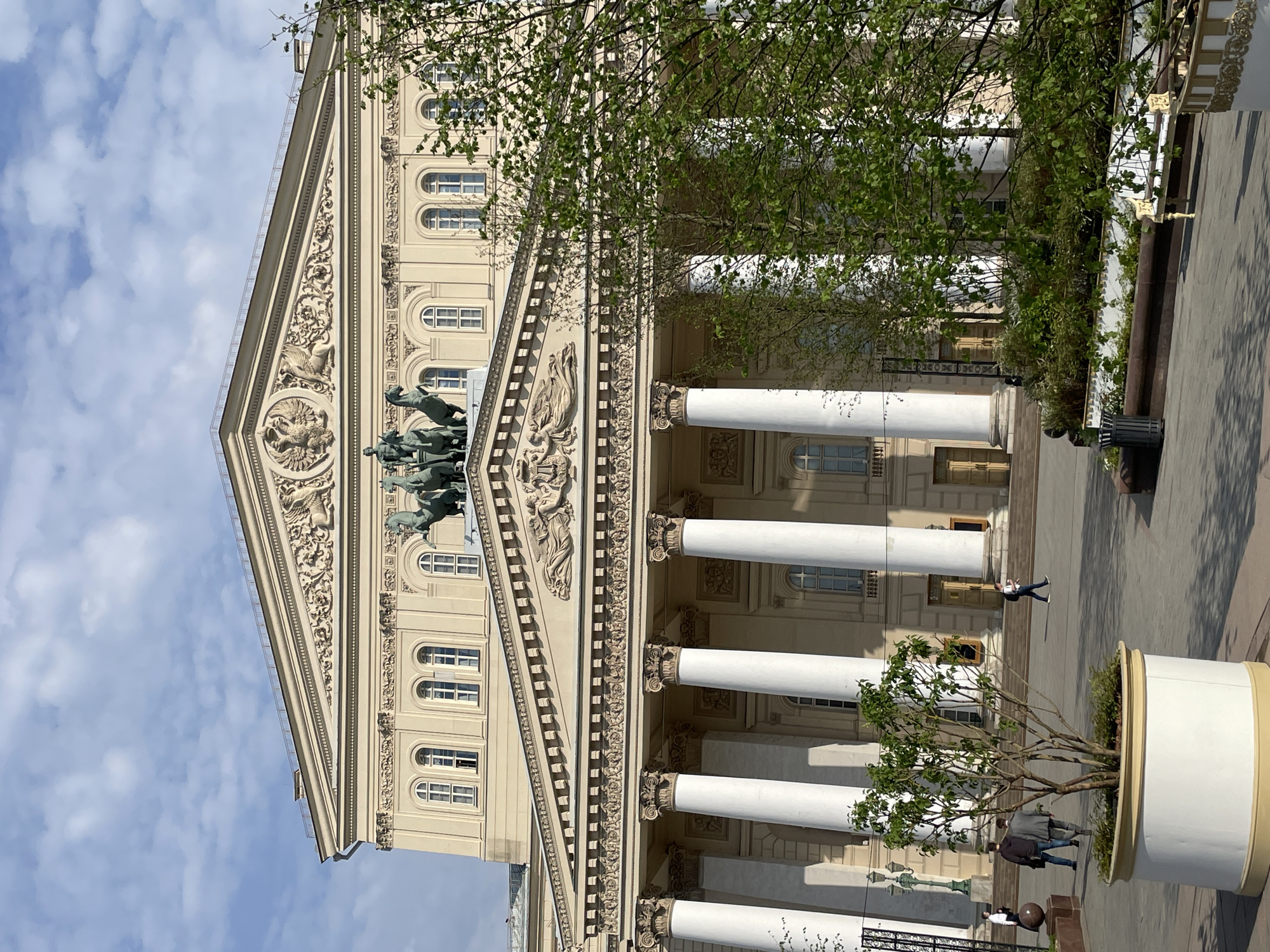 The image depicts the Bolshoi Theatre in Moscow, Russia, oriented in a portrait perspective. The theater's facade is prominently featured, showcasing its architectural details, including classical columns, ornate sculptures, and a quadriga sculpture atop the building's pediment.
Several people are visible in the foreground and near the building, some walking and one person is lying on the ground. A large potted tree stands in the foreground on the right side of the image. The sky is visible in the background, featuring a blue color and sparse clouds. The scene appears to be captured during the daytime under natural lighting conditions.