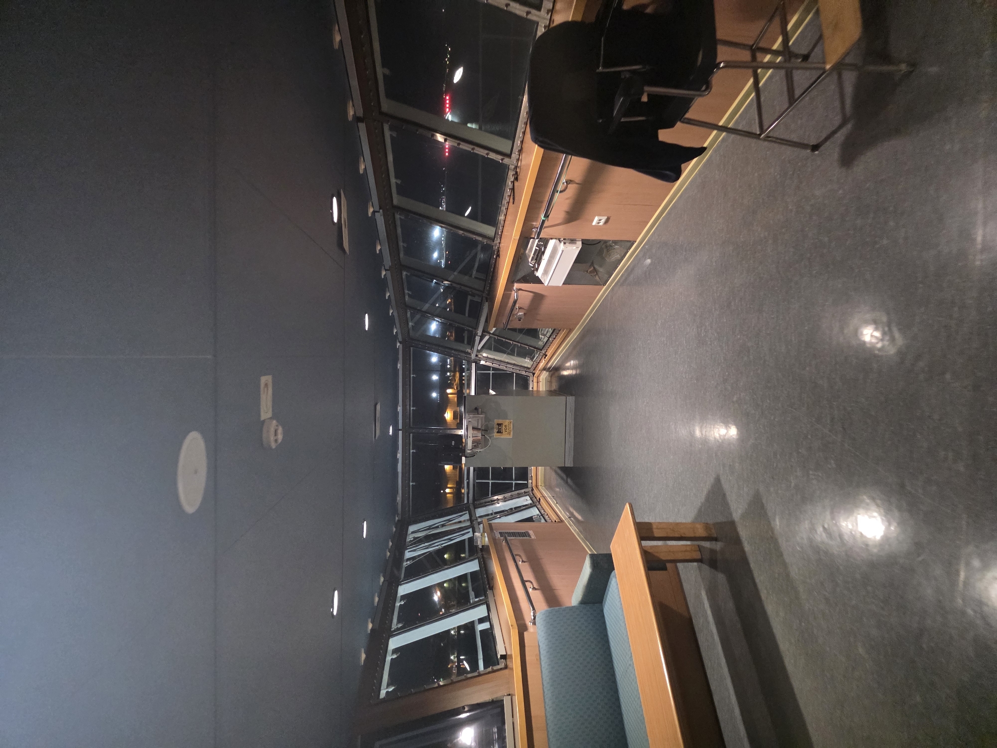 The image depicts the interior of a ship's bridge at night. The room has large windows offering a view of the port lights and water. A teal sofa with a wooden coffee table is on the left. In the center, a gray console with the label "VDR" is visible. On the right, a black captain's chair with a dark jacket draped over it is positioned near the windows. The floor is dark gray and reflective. The ceiling is a dark blue material with several recessed lights and a circular speaker. The scene appears to be located on a ferry terminal in Brindisi, Italy, given the presence of port infrastructure and vessels visible through the windows.