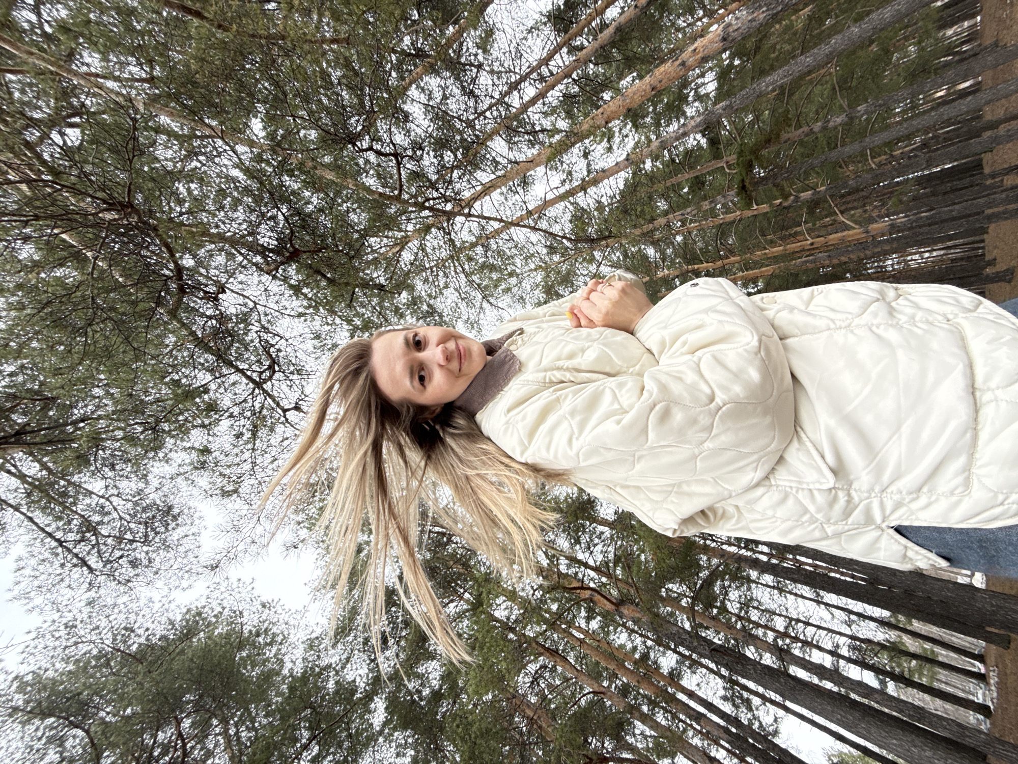 A young woman in a quilted white jacket stands in a pine forest in Chaikovsky, Russia. The perspective is from below, looking upwards, with tall pine trees surrounding her. The sky is overcast. Her blonde hair is blowing in the wind. She has her hands clasped together near her chest.