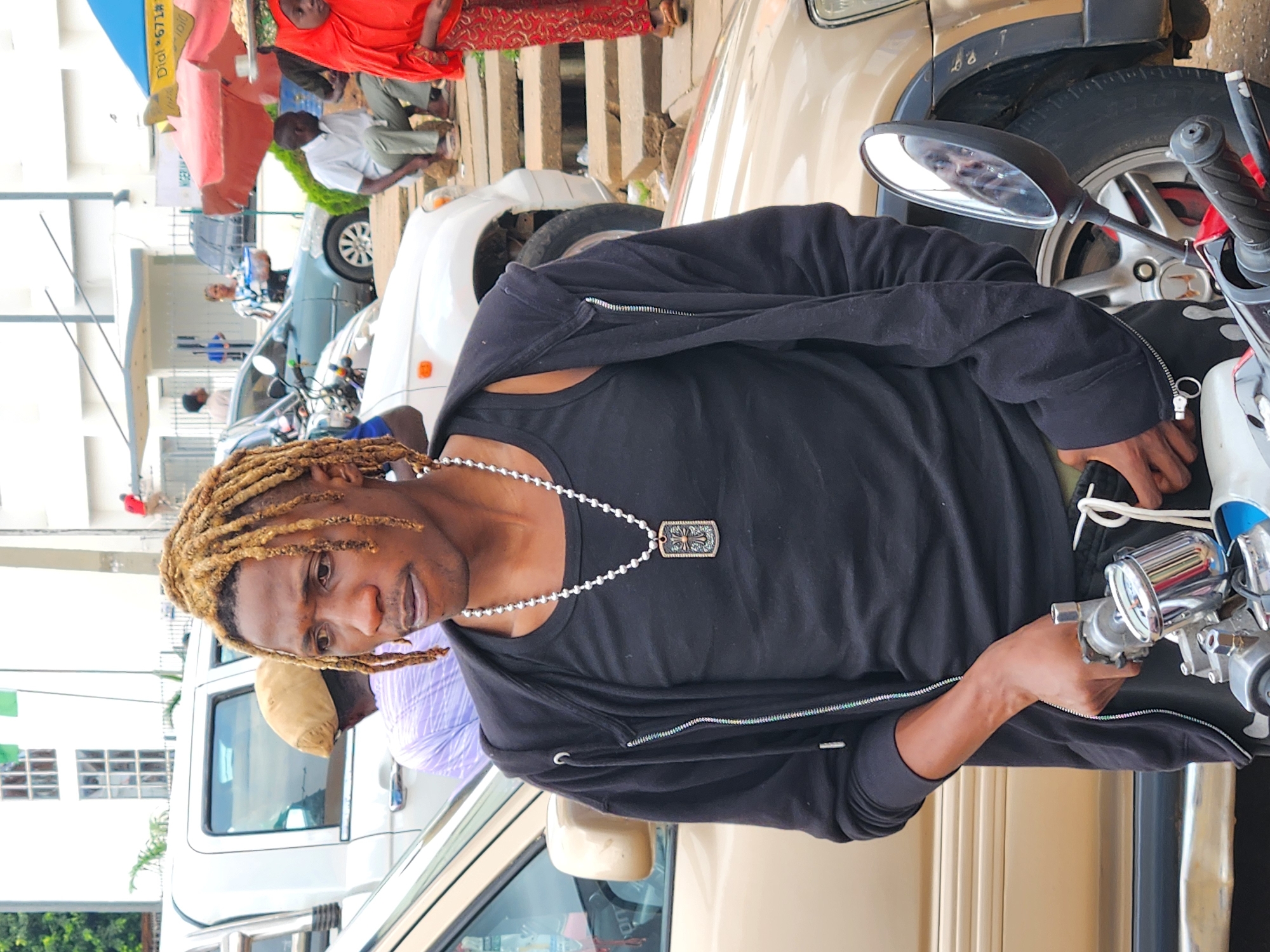 A medium shot captures a Black man with bleached dreadlocks standing next to a beige car. He is wearing a black tank top underneath an unzipped black hoodie and black pants. A silver chain with a rectangular pendant hangs around his neck. His right hand is resting on the handlebars of a motorcycle, which is partially visible in the foreground. In the background, other people are seen near cars and market stalls, with a white building visible behind them. The scene appears to be an outdoor street environment.