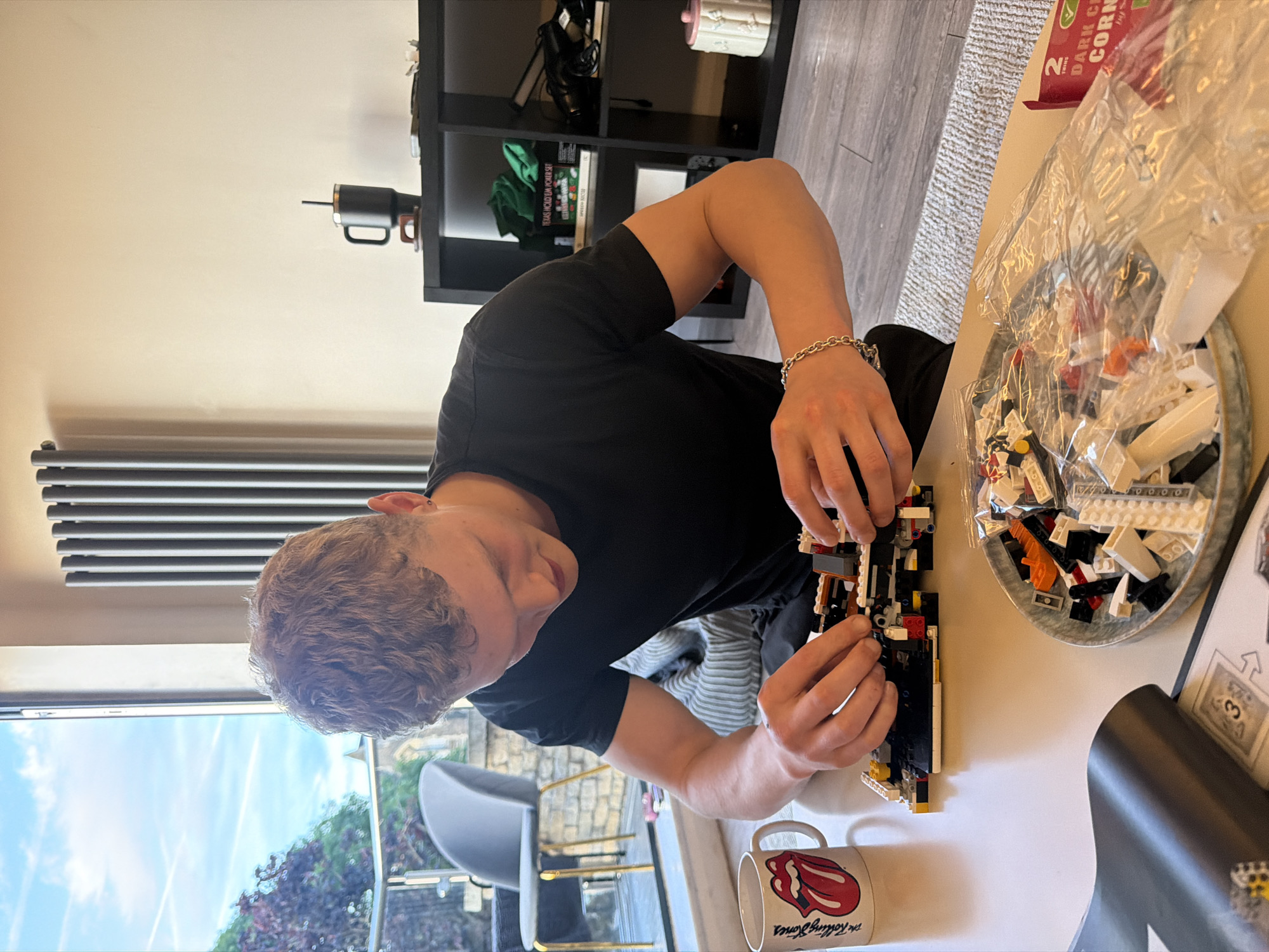 A person is sitting at a table assembling a Lego set. The person is wearing a black shirt and has a silver bracelet on their wrist. The table is white and has a Rolling Stones mug on it. There is a bowl of Lego pieces and the instruction manual on the table. There is a shelf in the background with various objects on it, including a coffee cup and a Texas Hold 'Em poker set. Outside the window, the sky is blue.
It is not possible to discern the location is in London, United Kingdom.
