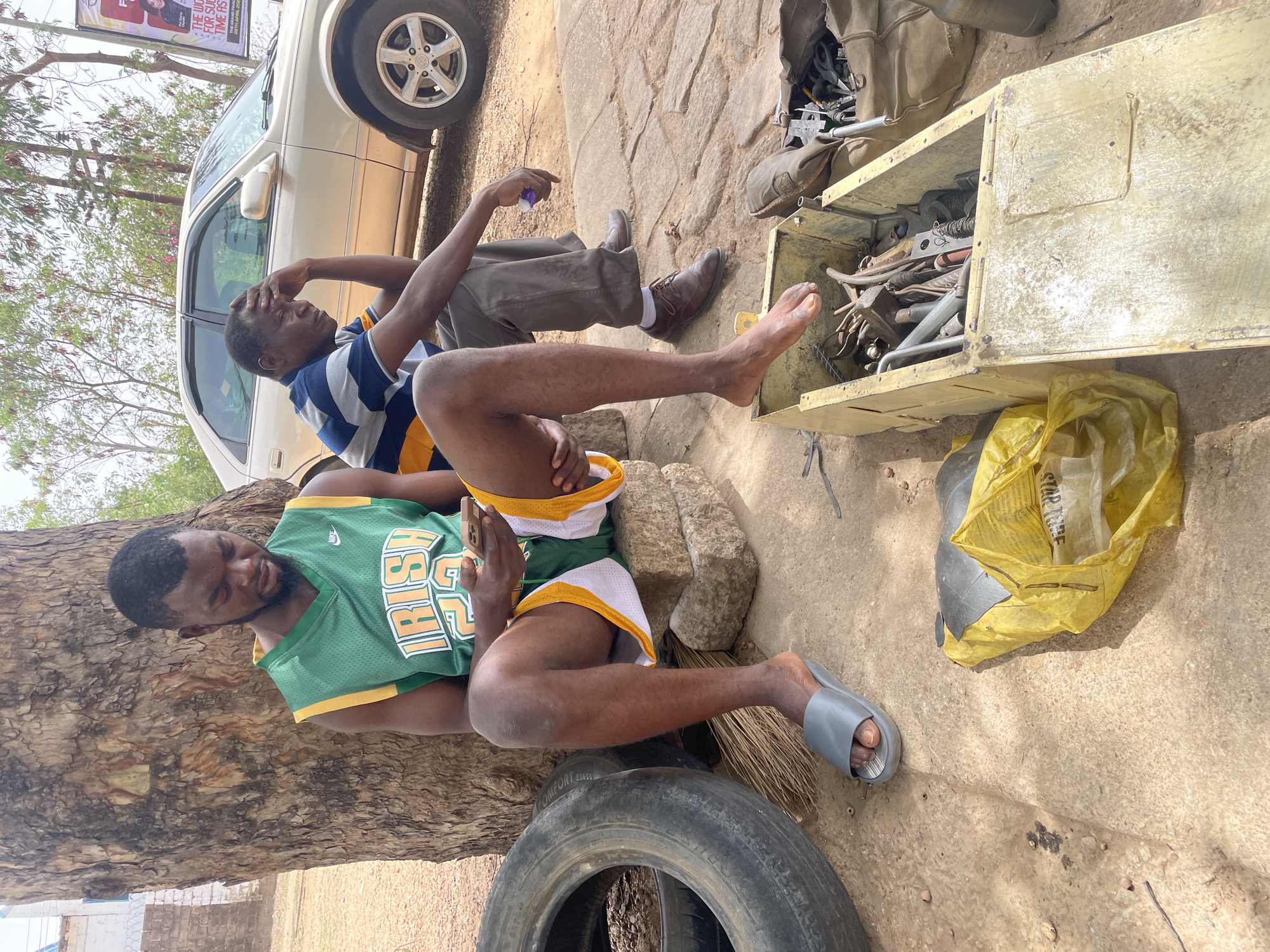 The scene is an outdoor setting in the city of Jos, Nigeria. Two men are seated or reclining on the ground, next to a car. One man, wearing a green and yellow "IRISH 25" basketball jersey and grey slides, is leaning against a tree trunk while using a mobile phone. The second man, wearing a striped shirt and brown shoes, is sitting with his hand on his forehead, appearing pensive.
Next to them is a weathered, open metal toolbox filled with metal objects. A yellow plastic bag sits nearby. A tire lies on the ground next to the man in the jersey. The background includes a light-colored car, a sign partially visible in the upper left corner, and sparse foliage. The ground is a dirt or concrete surface.